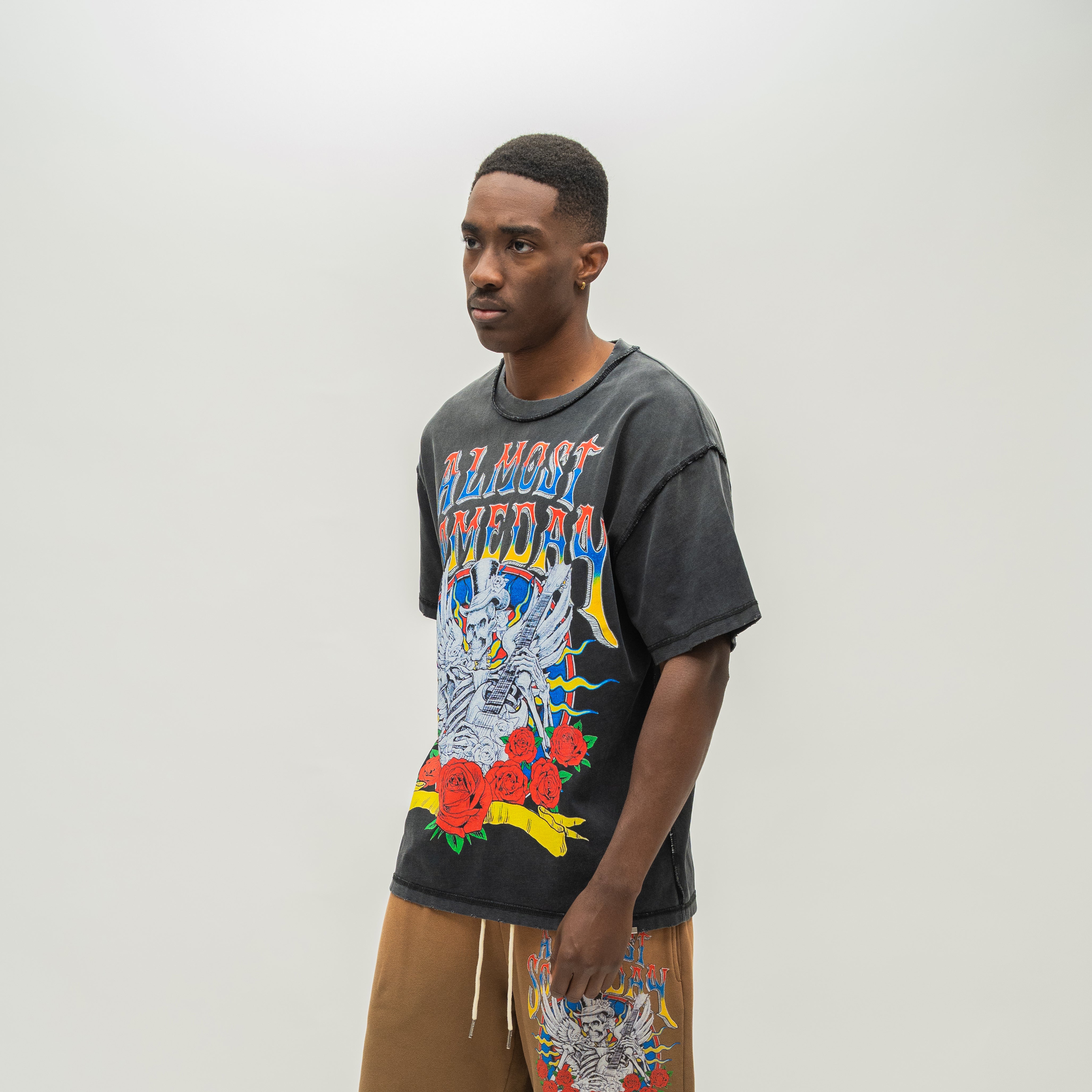 A man wearing a faded black Almost Someday graphic t-shirt with a colorful skeleton and rose design, paired with brown drawstring shorts, standing against a light gray studio background.