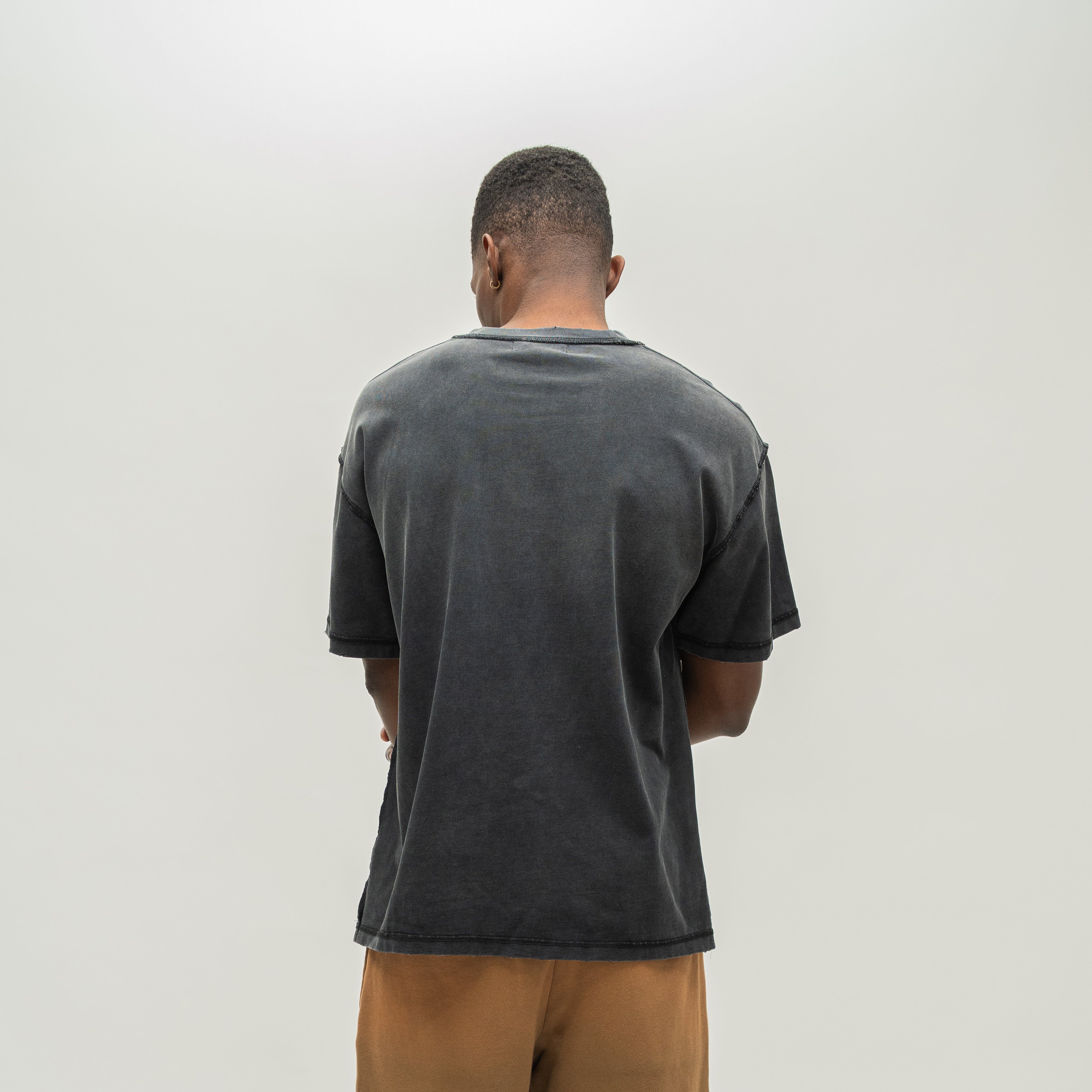 A man viewed from behind wearing a faded black washed t-shirt and tan pants, standing against a plain light gray background.