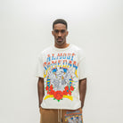 A man wearing a cream-colored graphic t-shirt with a colorful 'Almost Someday' skeleton illustration, paired with brown sweatshorts, against a plain white studio background.