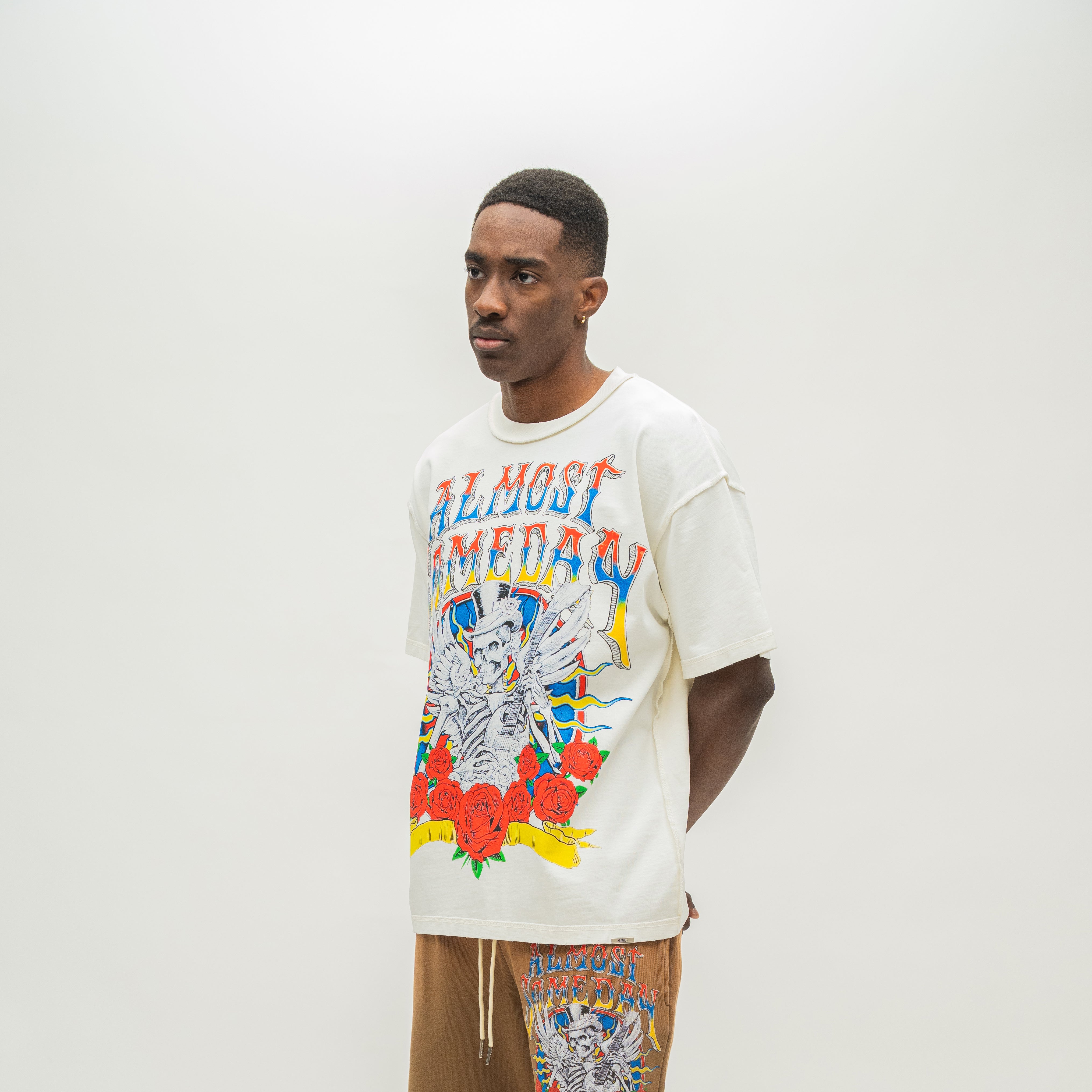A man wearing a cream-colored graphic t-shirt with a colorful skeleton and text design, paired with brown sweatshorts, standing against a plain light gray background.