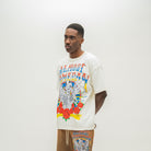 A man wearing a cream-colored graphic t-shirt with a colorful skeleton and text design, paired with brown sweatshorts, standing against a plain light gray background.