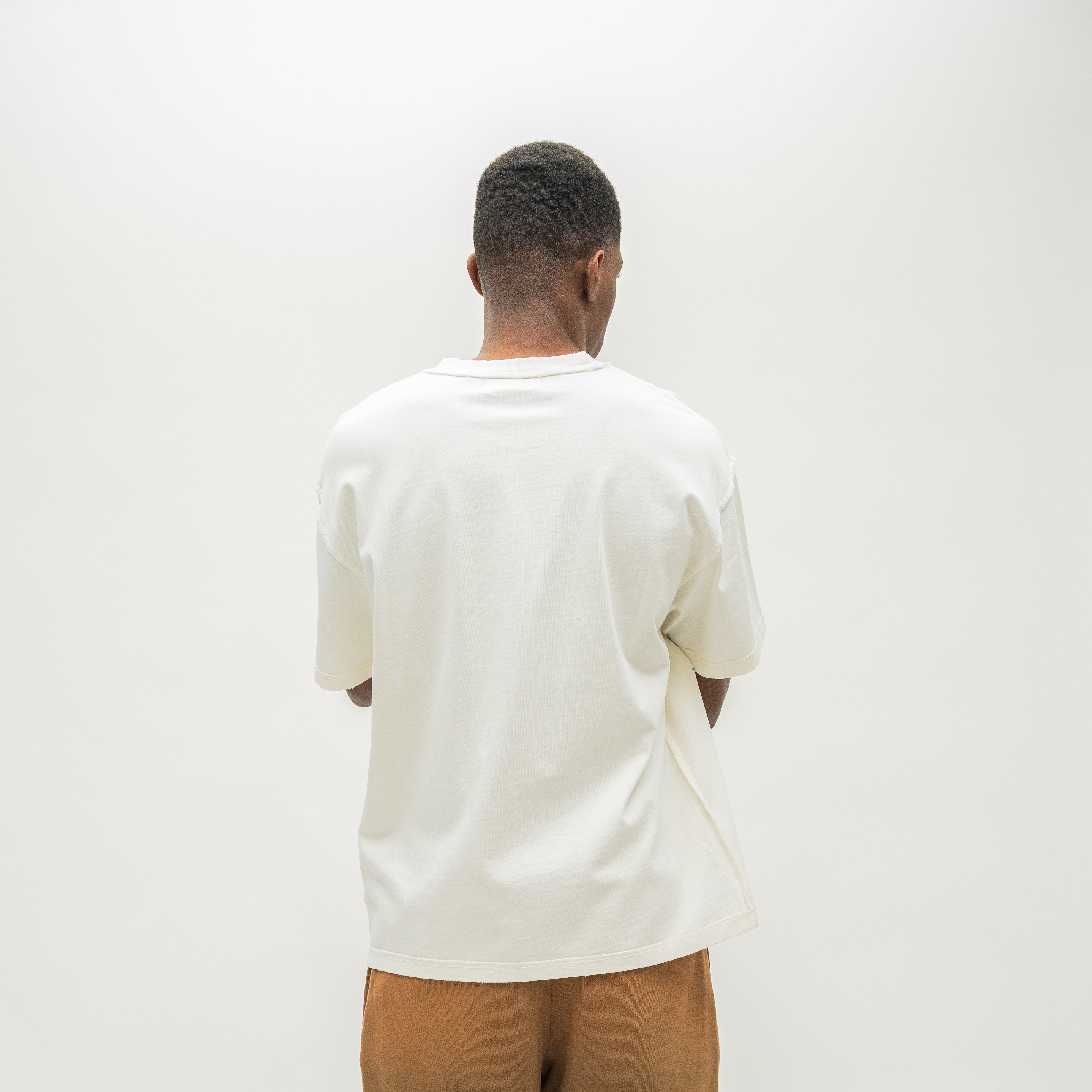 A man viewed from behind wearing an oversized off-white t-shirt and tan sweatshorts, standing against a plain white studio background.