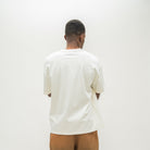 A man viewed from behind wearing an oversized off-white t-shirt and tan sweatshorts, standing against a plain white studio background.