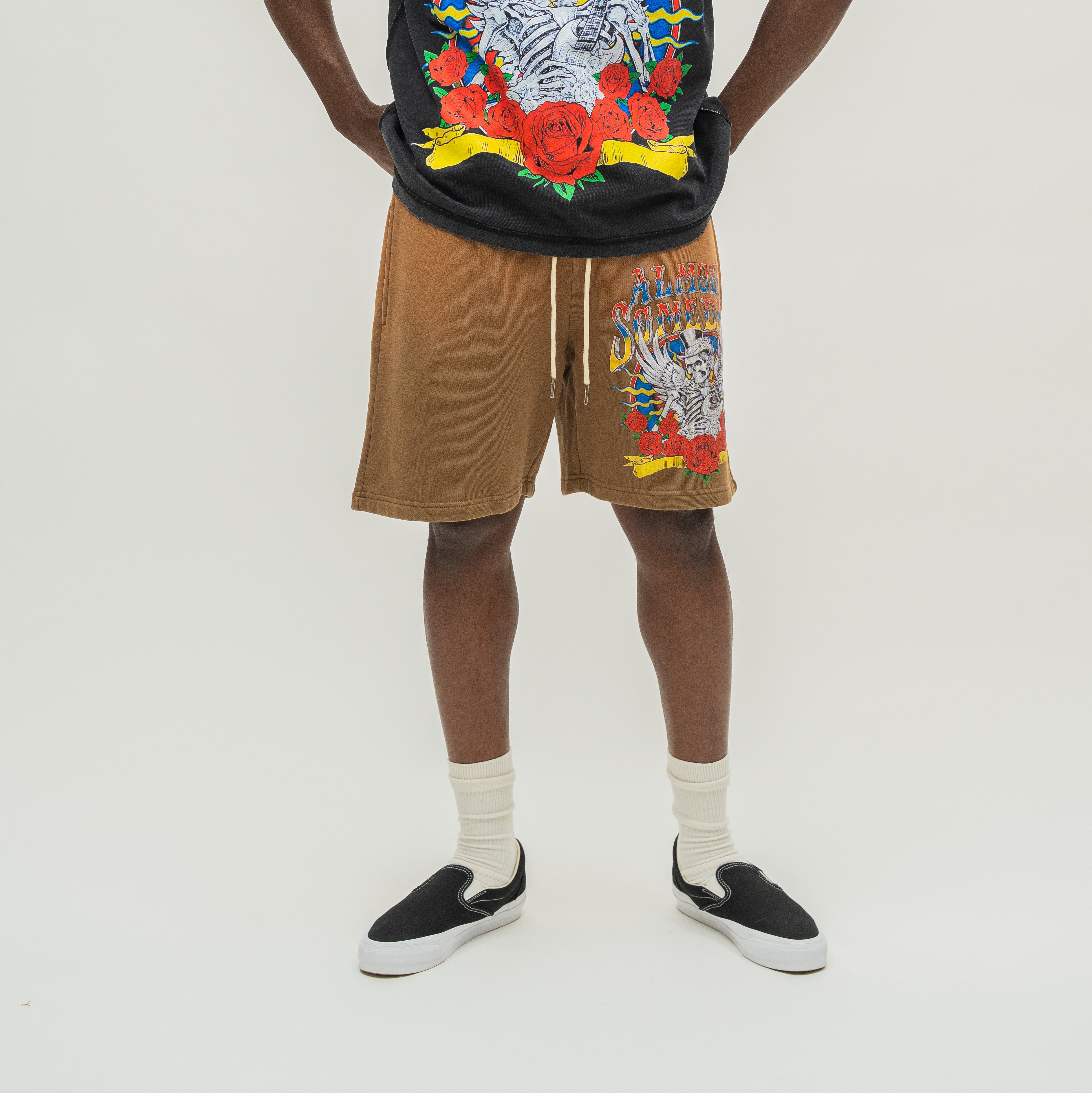 A person wearing tan drawstring shorts with a colorful skeleton graphic on the left leg, paired with a black graphic t-shirt, white socks, and black slip-on shoes, photographed against a plain light background.