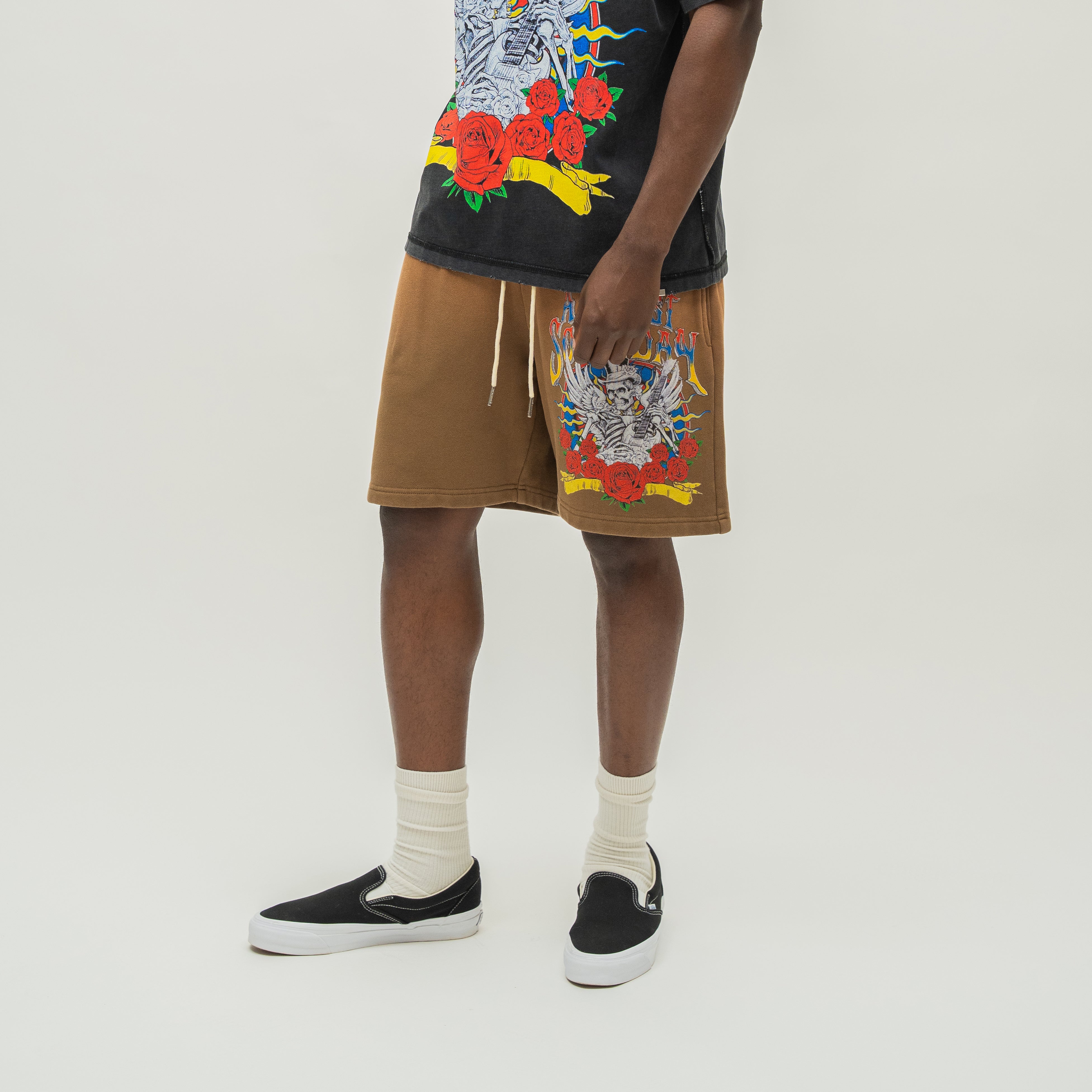A model wearing brown drawstring shorts with colorful skeleton and floral graphics on the front, paired with a black graphic t-shirt, white socks, and black slip-on sneakers, photographed against a light gray studio background.