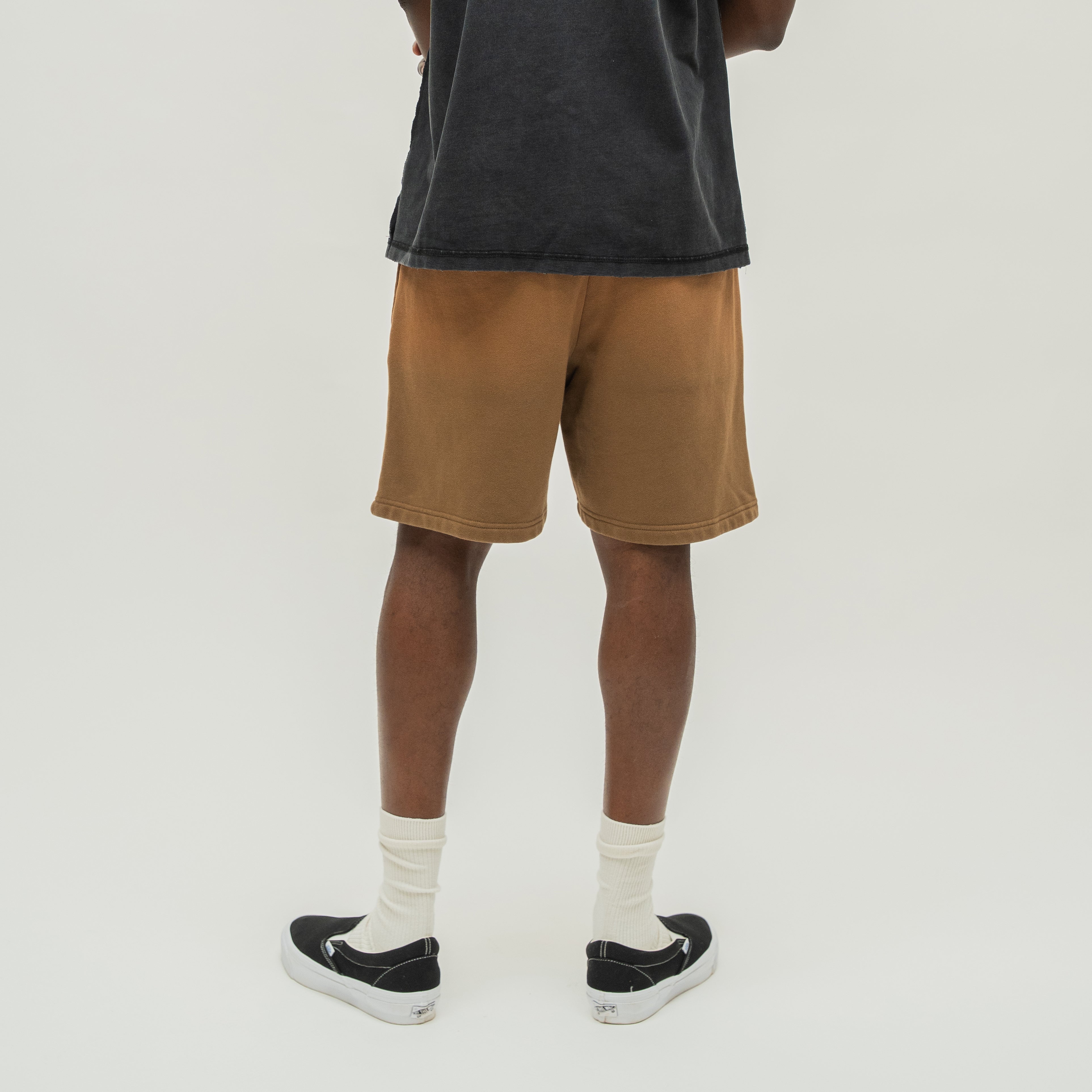 A person shown from the back wearing brown washed denim shorts, a dark charcoal t-shirt, white socks, and black slip-on sneakers against a light gray studio background.
