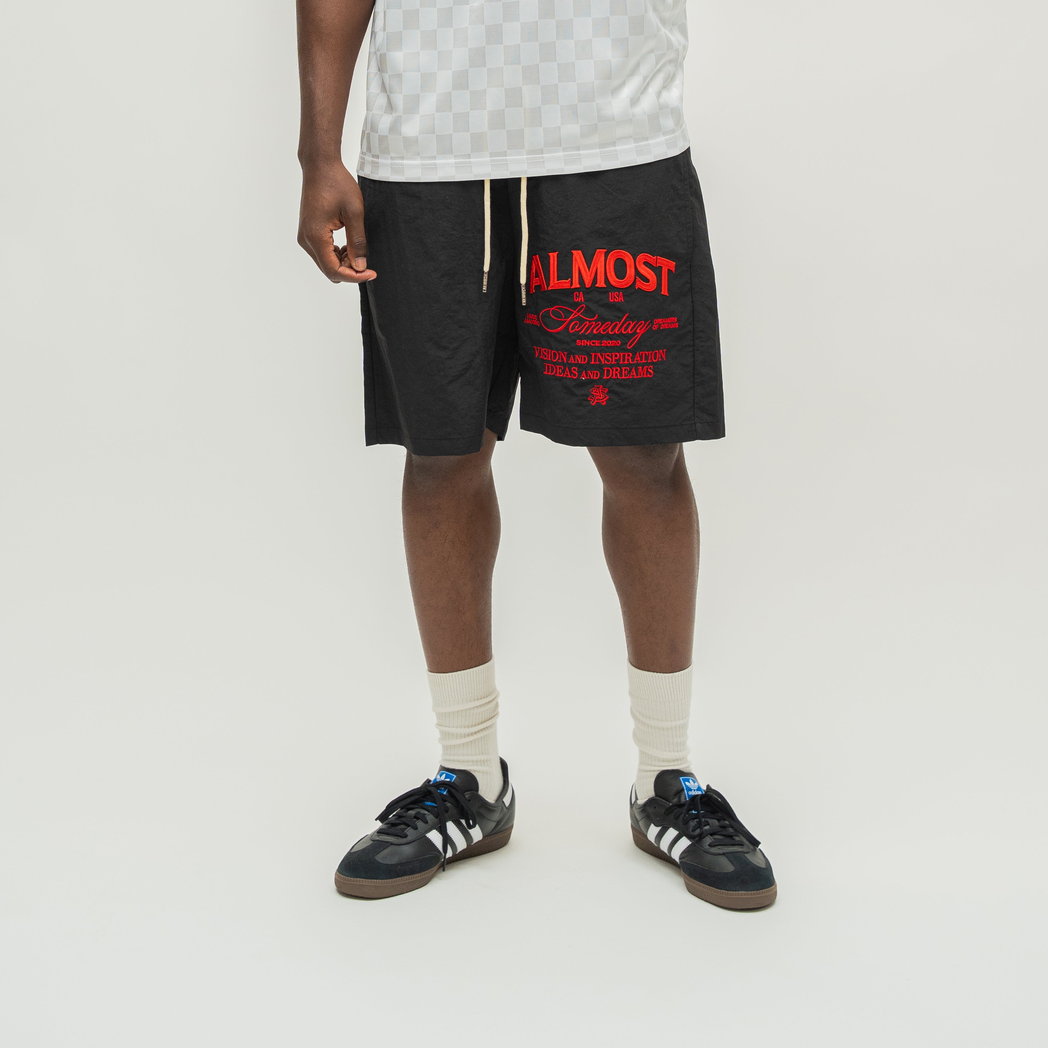 A model from the waist down wearing black athletic shorts with red and white graphic text on the left leg, paired with a white patterned tank top, white socks, and black sneakers.