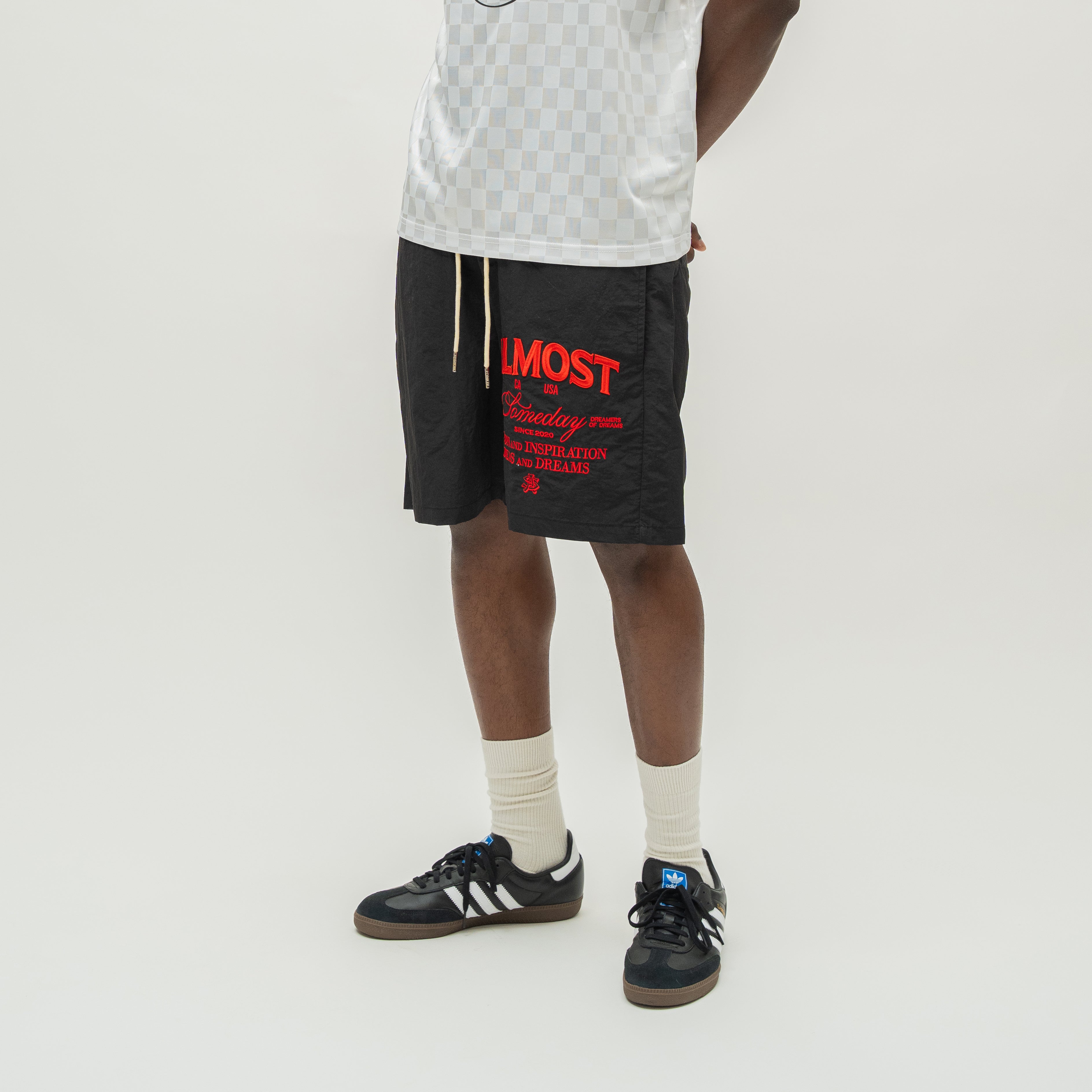 A model from the waist down wearing black athletic shorts with red and white text, paired with a white patterned tank top, off-white socks, and black sneakers with white stripes, against a plain light gray background.