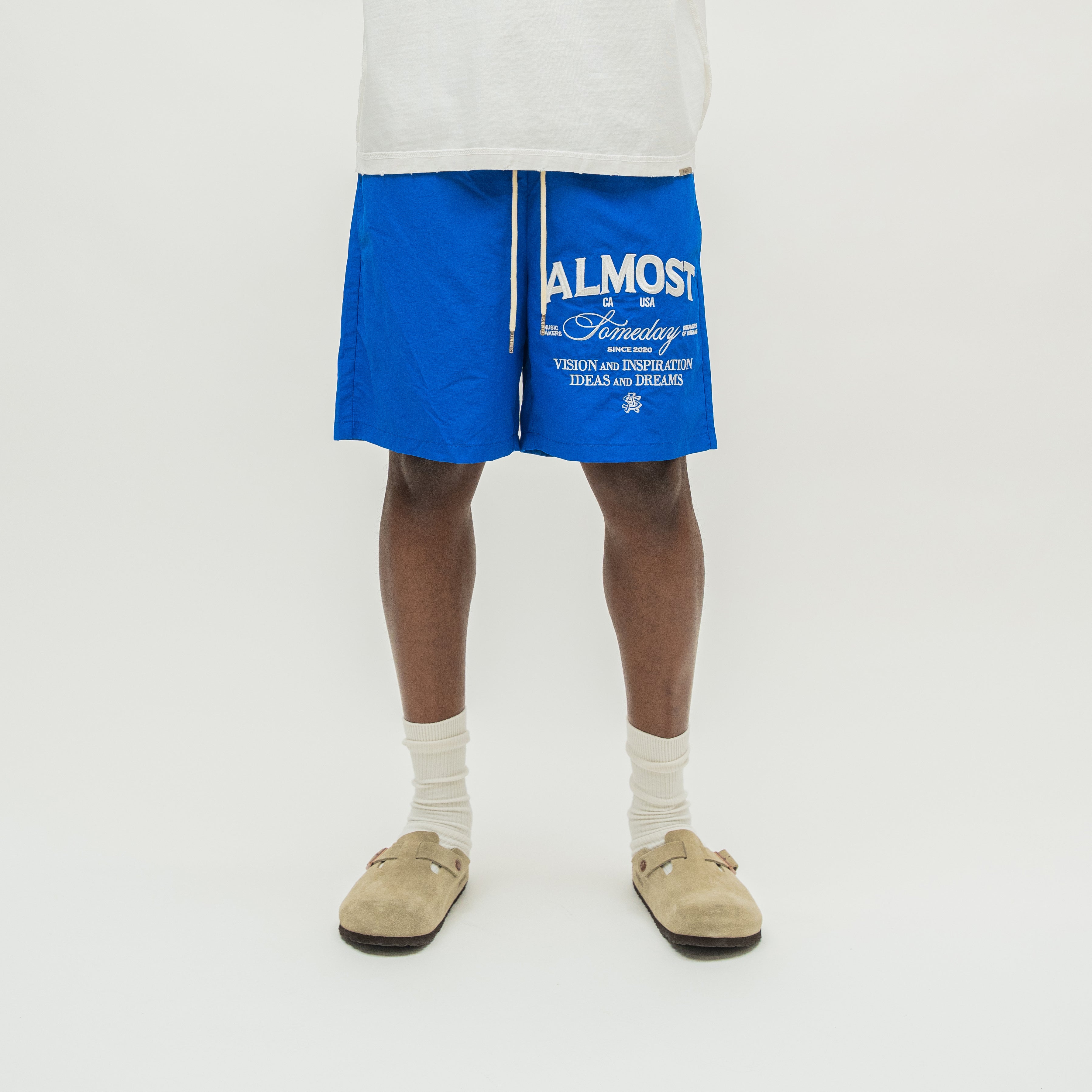 A person from the waist down wearing bright blue drawstring shorts with white text, white socks, and beige slip-on shoes against a plain light background.