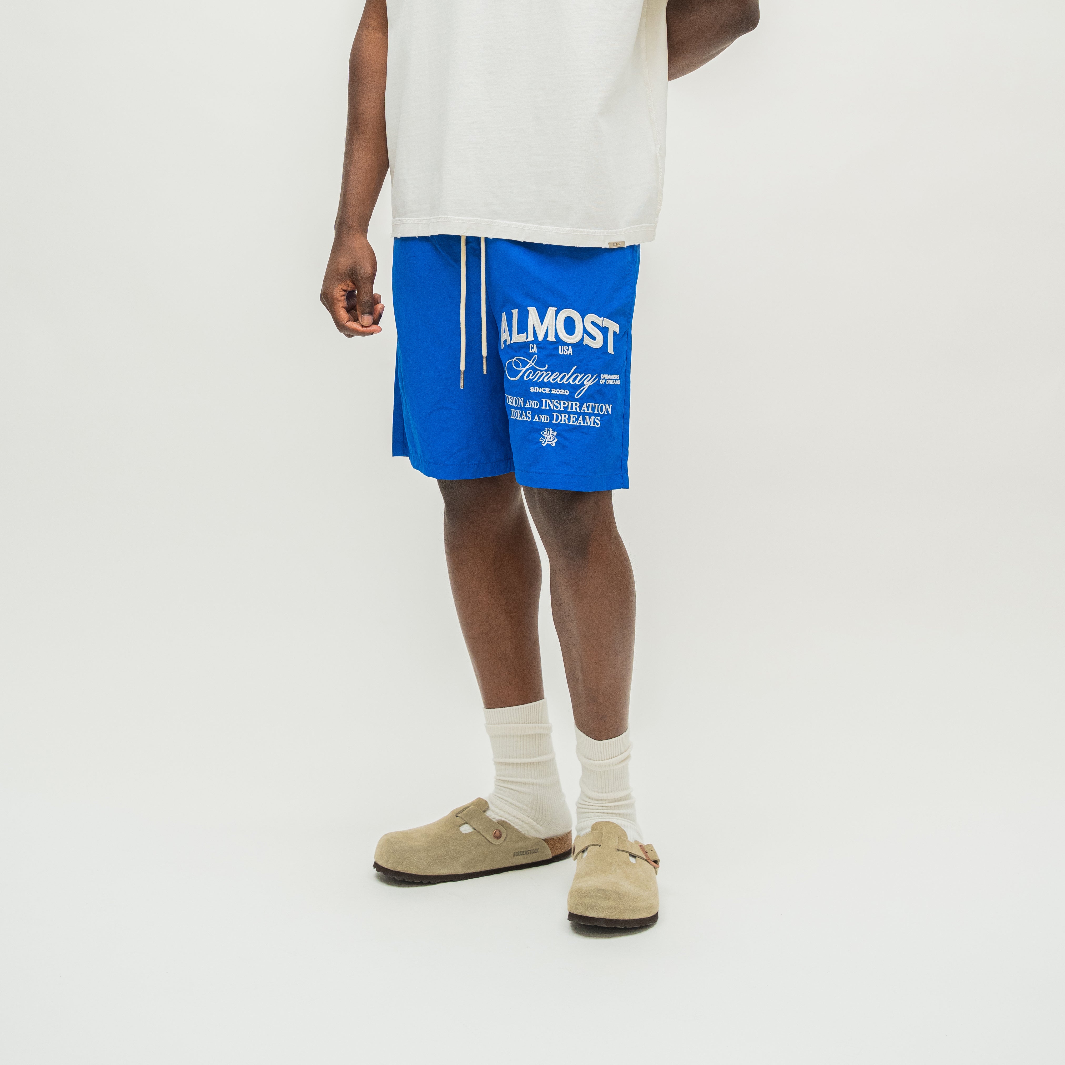 A model from the waist down wearing bright blue drawstring shorts with white graphic text, a white t-shirt, white socks, and beige clogs, photographed against a clean white studio background.