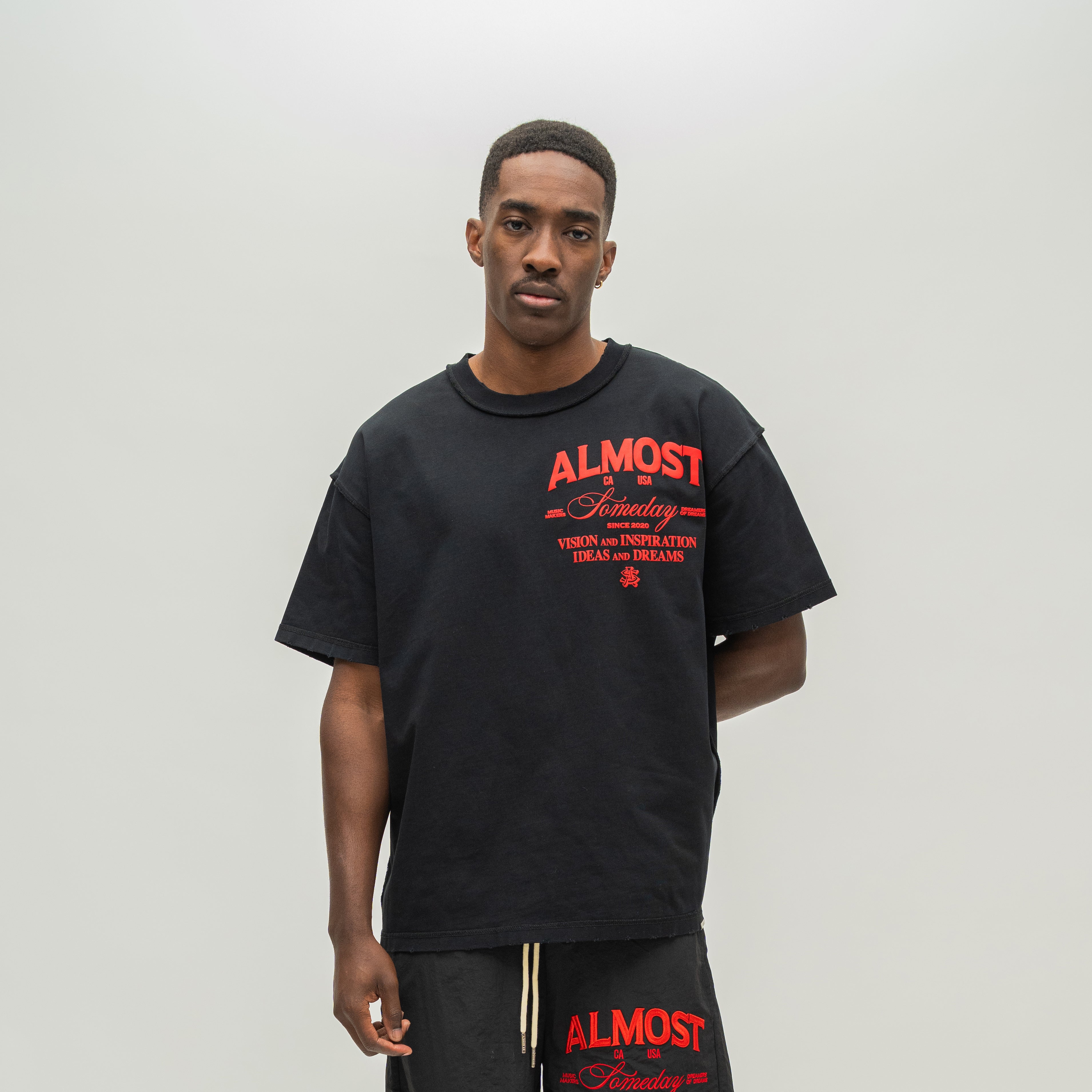 A man wearing a black oversized t-shirt and matching sweatpants with red graphic text on the front, standing against a plain light gray studio background.