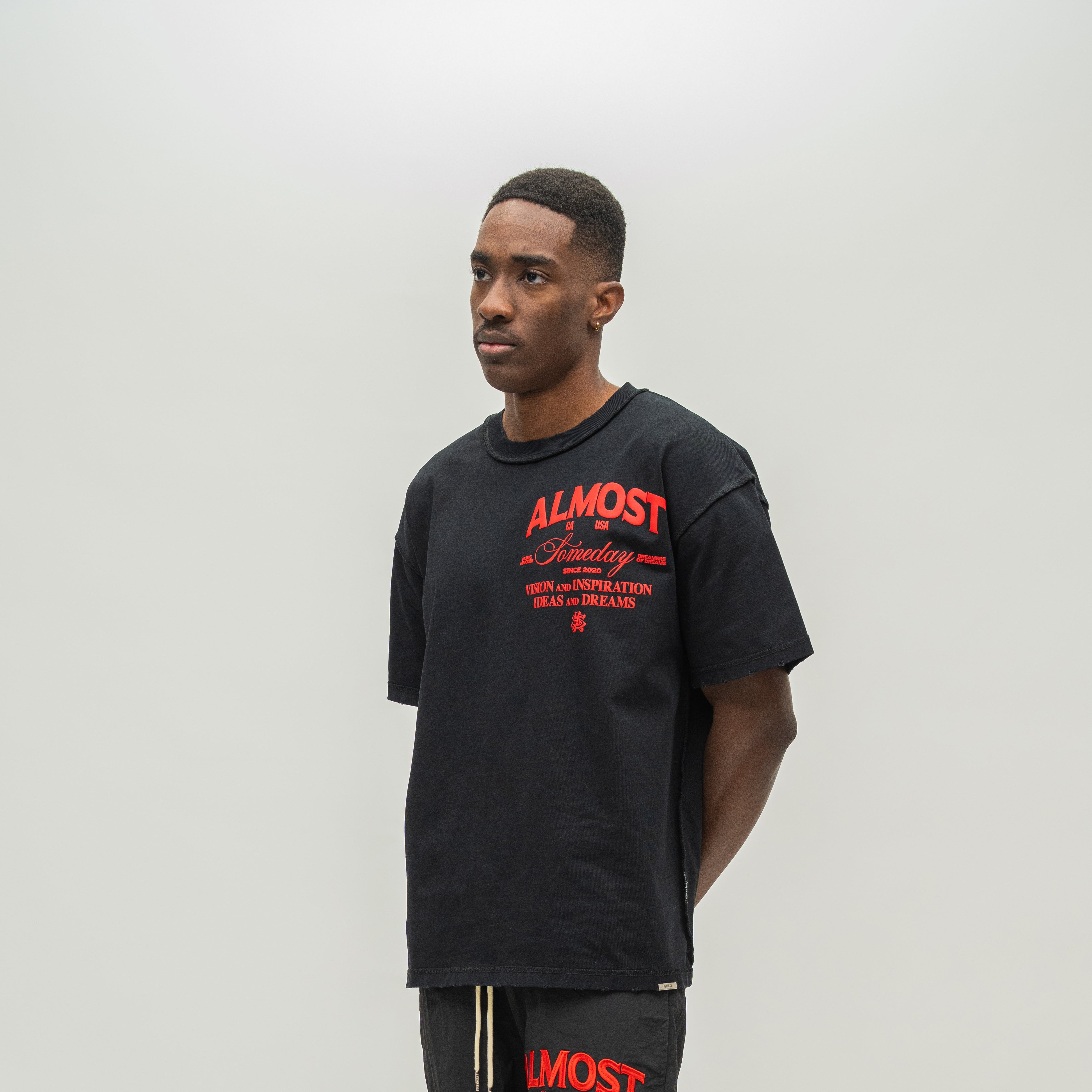 A man wearing a black oversized t-shirt with red graphic text on the front, standing against a plain light gray studio background.