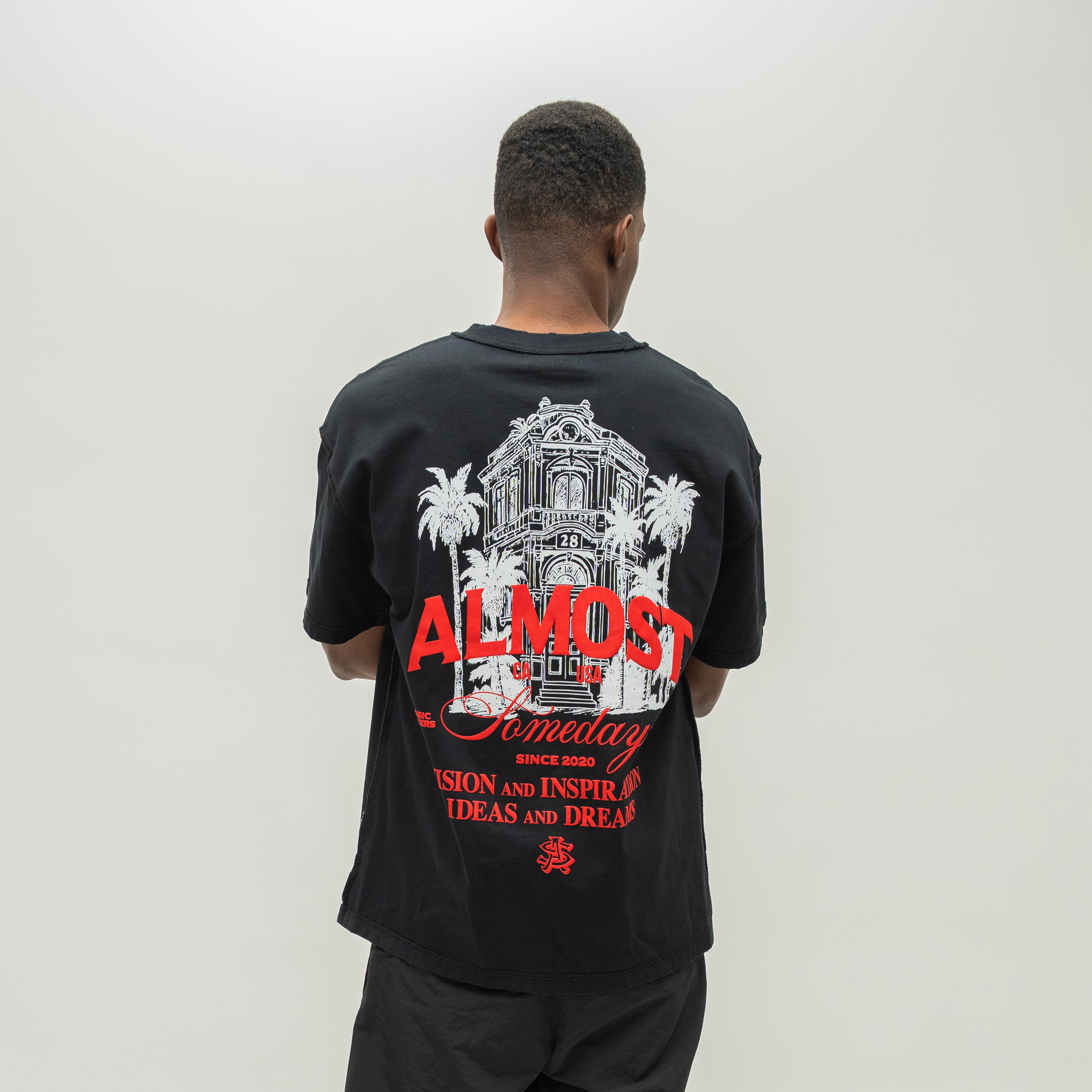A man viewed from behind wearing a black t-shirt with a large graphic print on the back. The graphic features a white architectural illustration of a cathedral flanked by palm trees, with the words 'ALMOST Someday' in red and white text.
