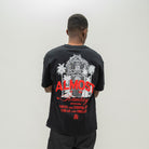 A man viewed from behind wearing a black t-shirt with a large graphic print on the back. The graphic features a white architectural illustration of a cathedral flanked by palm trees, with the words 'ALMOST Someday' in red and white text.