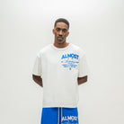 A man modeling an oversized off-white t-shirt with blue graphic text on the chest that reads 'ALMOST SOMEDAY, paired with blue shorts against a plain light gray background.