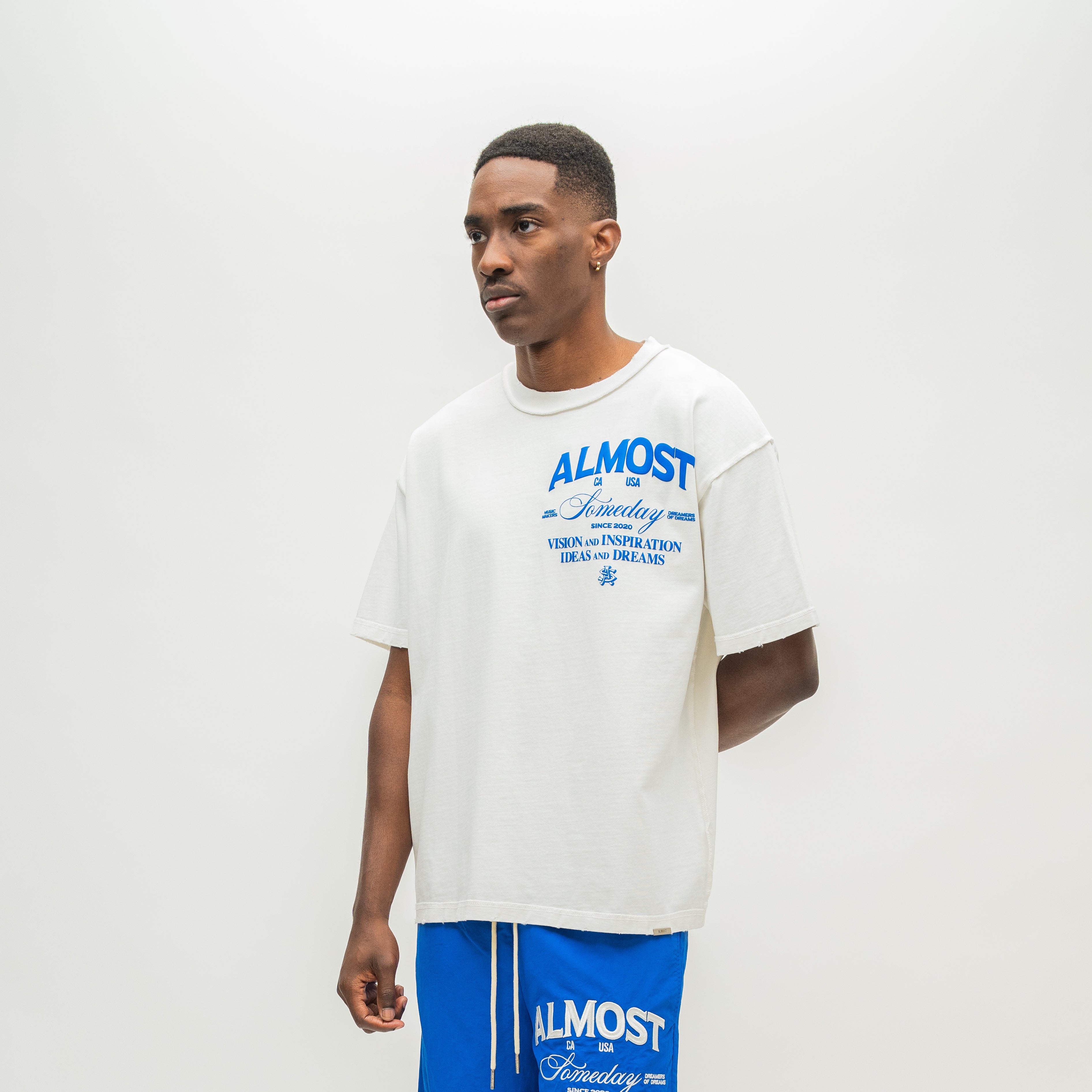 A man wearing an oversized white t-shirt with blue graphic text that reads 'ALMOST SOMEDAY' and blue athletic shorts with matching blue text, standing against a plain off-white studio background.