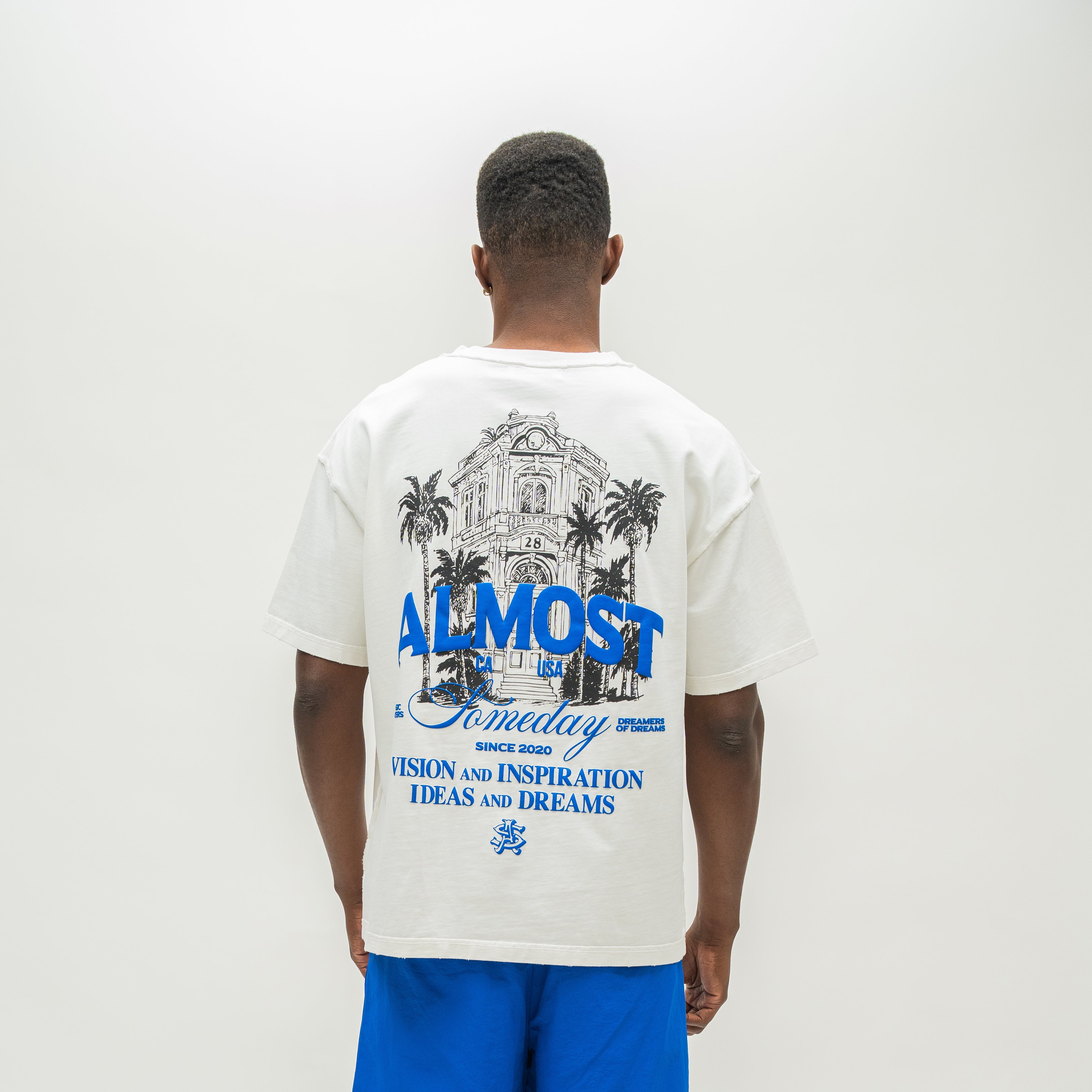 A man viewed from behind wearing an oversized off-white graphic t-shirt with a blue and black illustration and text on the back, paired with bright blue shorts, against a plain white studio background.