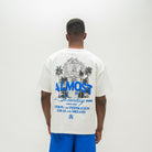 A man viewed from behind wearing an oversized off-white graphic t-shirt with a blue and black illustration and text on the back, paired with bright blue shorts, against a plain white studio background.