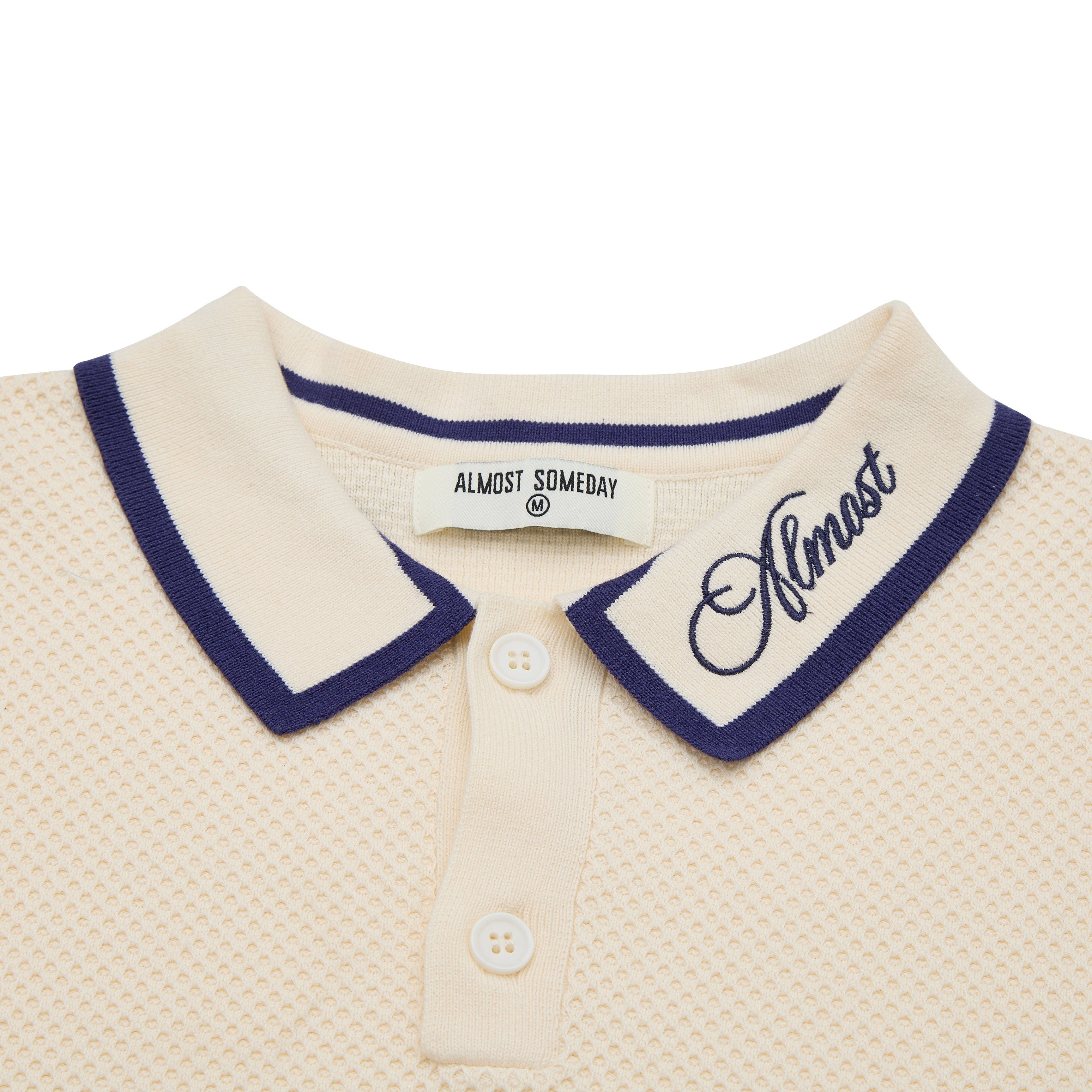 Close-up of a cream-colored polo shirt with navy trim and 'Almost' branding.