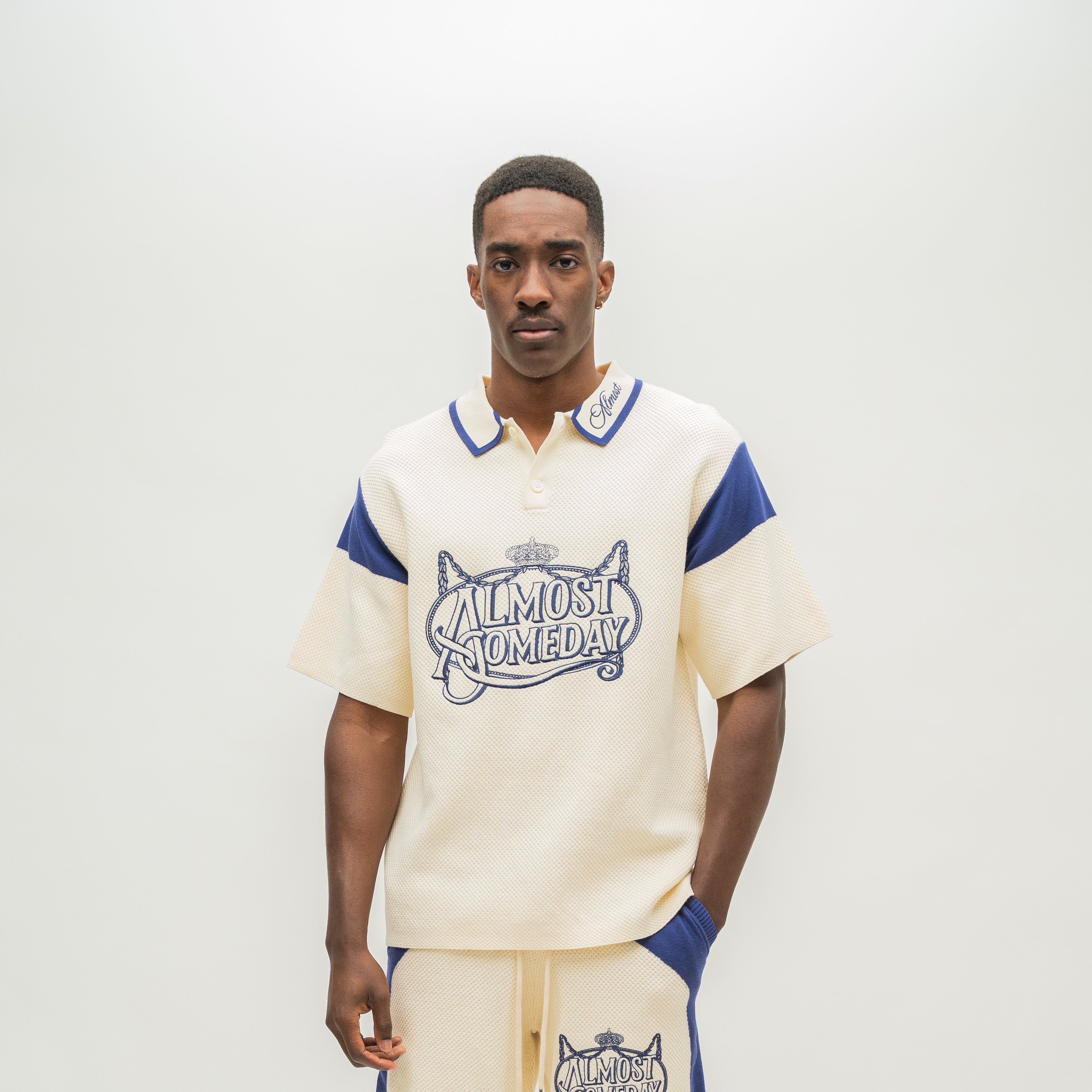 A man modeling a cream-colored short-sleeve polo shirt with blue accents and a graphic reading 'ALMOST SOMEDAY' on the front, paired with matching cream pants with blue side panels, against a plain light background.