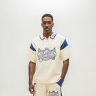 A man modeling a cream-colored short-sleeve polo shirt with blue accents and a graphic reading 'ALMOST SOMEDAY' on the front, paired with matching cream pants with blue side panels, against a plain light background.