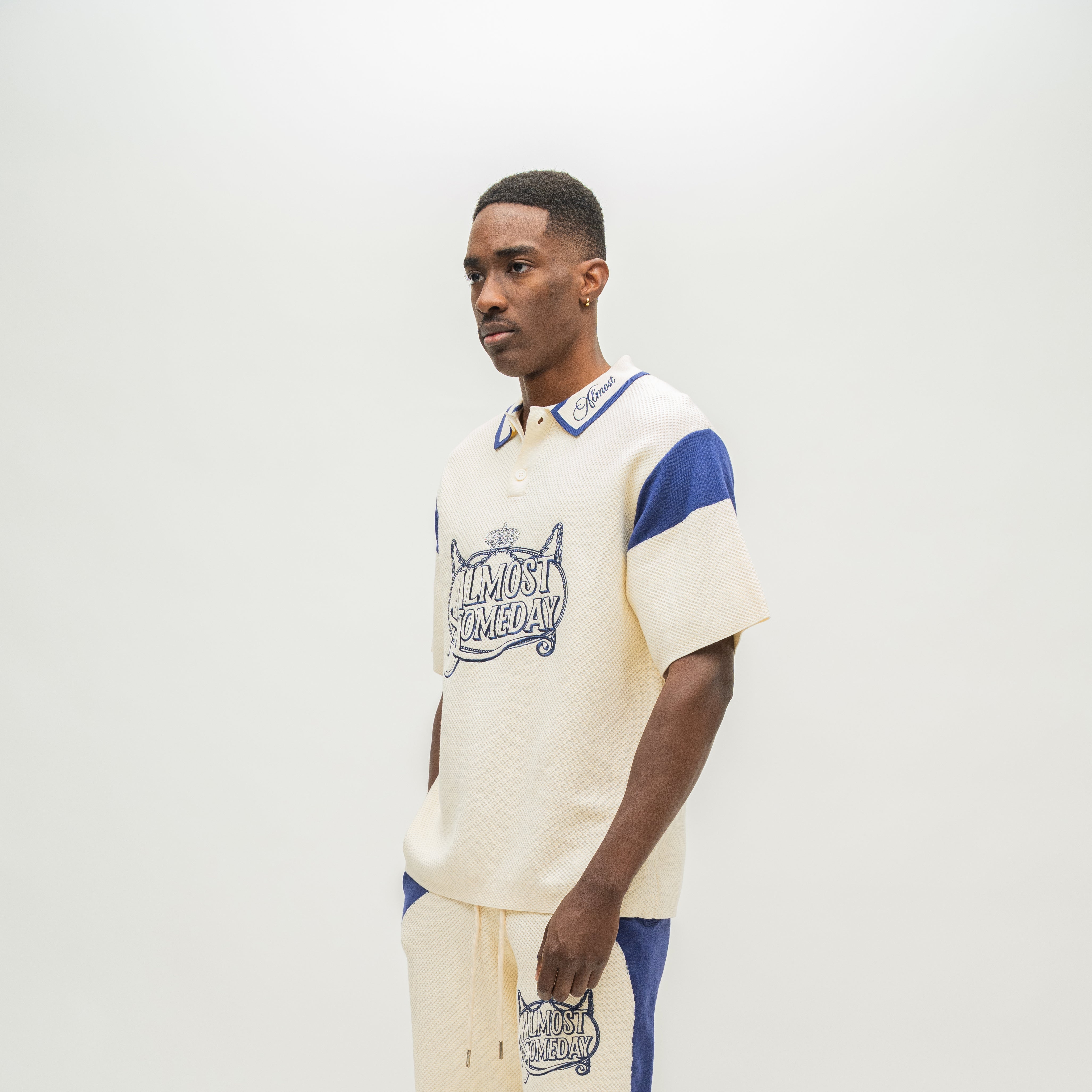 A man modeling a cream-colored short-sleeve polo shirt with blue accents and a graphic print on the front, paired with matching cream pants with blue side panels, against a plain light gray background.