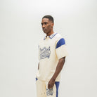 A man modeling a cream-colored short-sleeve polo shirt with blue accents and a graphic print on the front, paired with matching cream pants with blue side panels, against a plain light gray background.