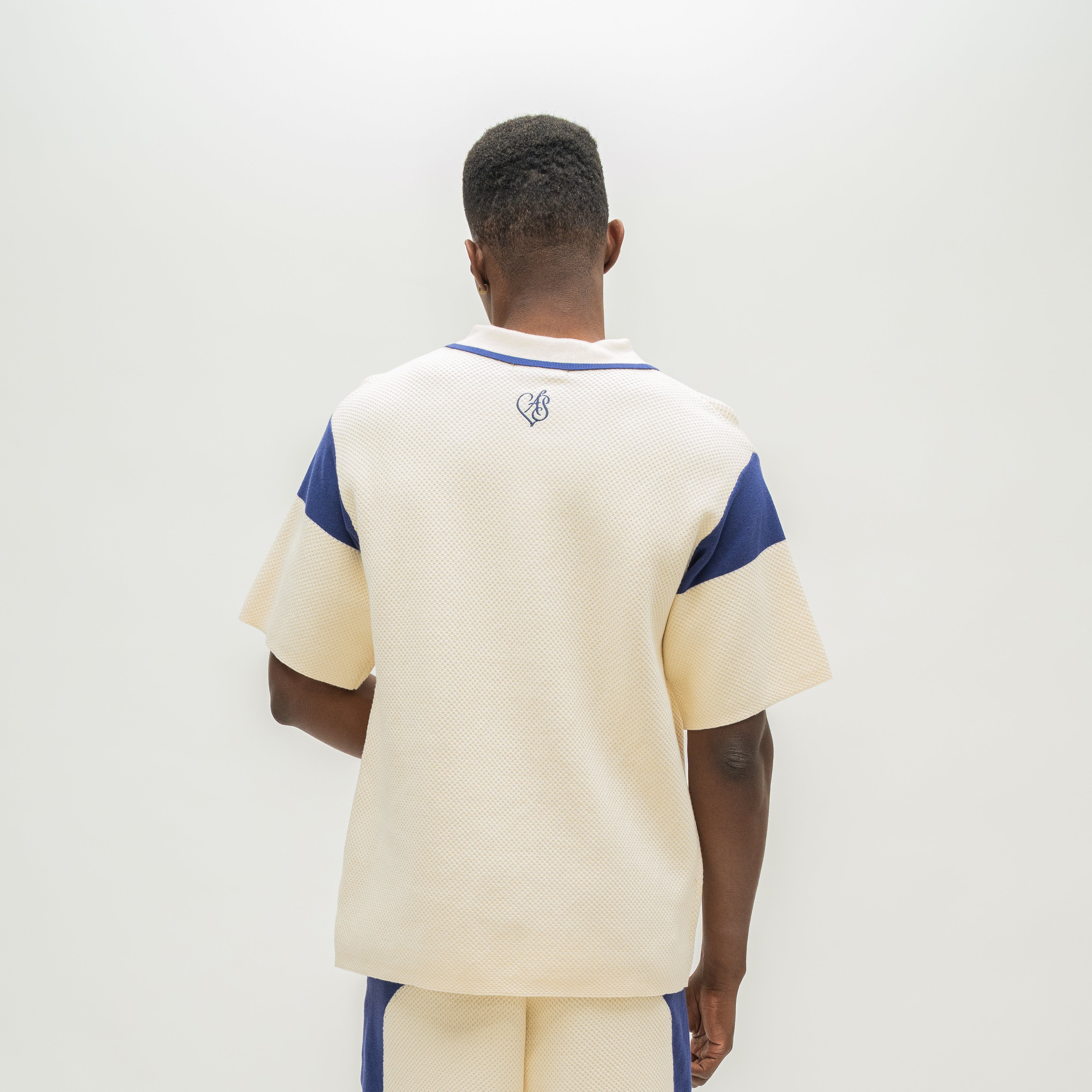 A man viewed from behind wearing a cream-colored short-sleeve knit top with blue shoulder panels and a small blue chest logo, paired with matching cream and blue shorts, against a plain white background.