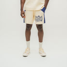 A model is shown from the waist down wearing cream-colored shorts with blue side panels and a navy graphic print on the left leg. The shorts feature a drawstring waist and are paired with cream socks and cream sneakers on a light gray studio background.
