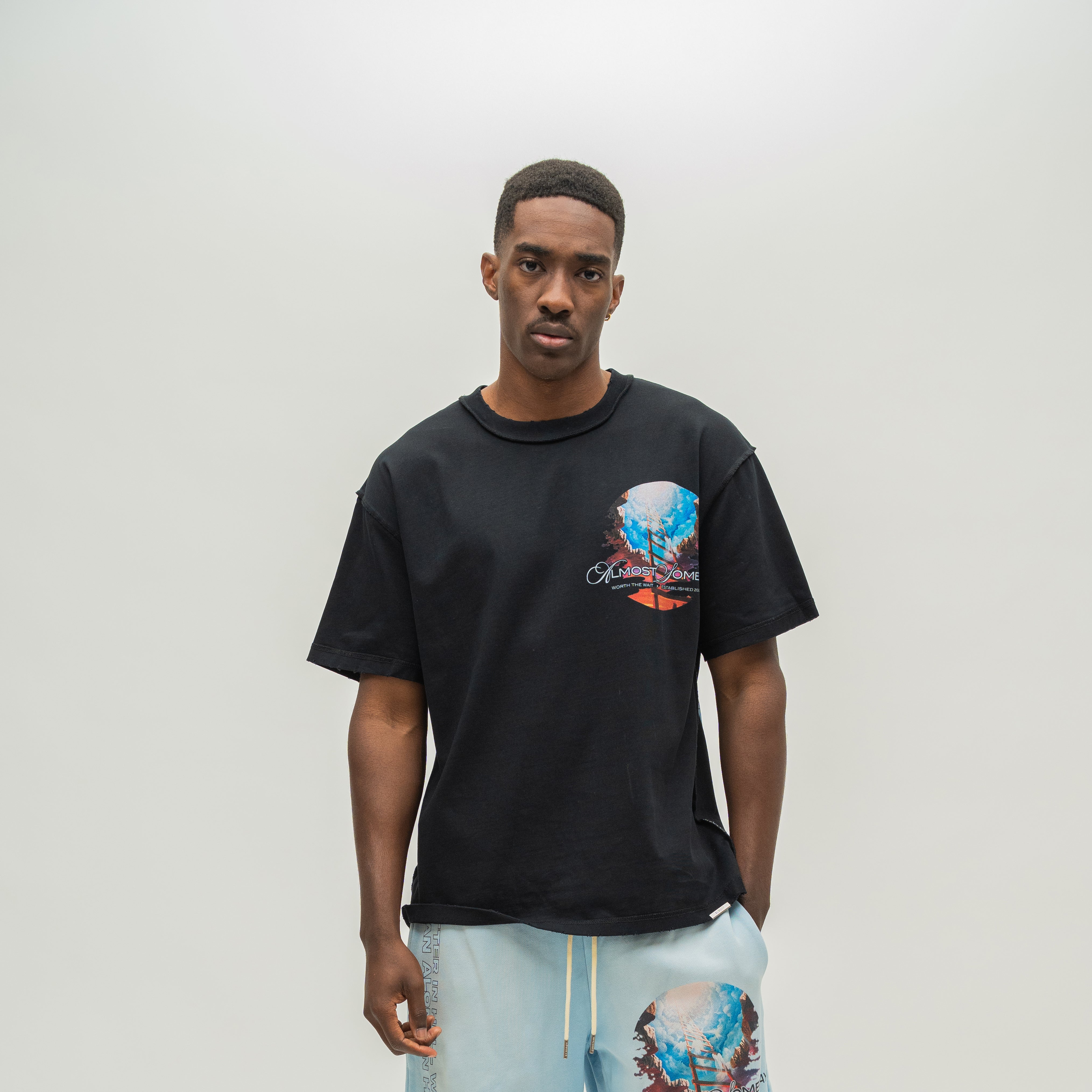 A man wearing a black short-sleeve graphic t-shirt with a colorful abstract print on the left chest, paired with light blue drawstring shorts, standing against a plain light gray studio background.