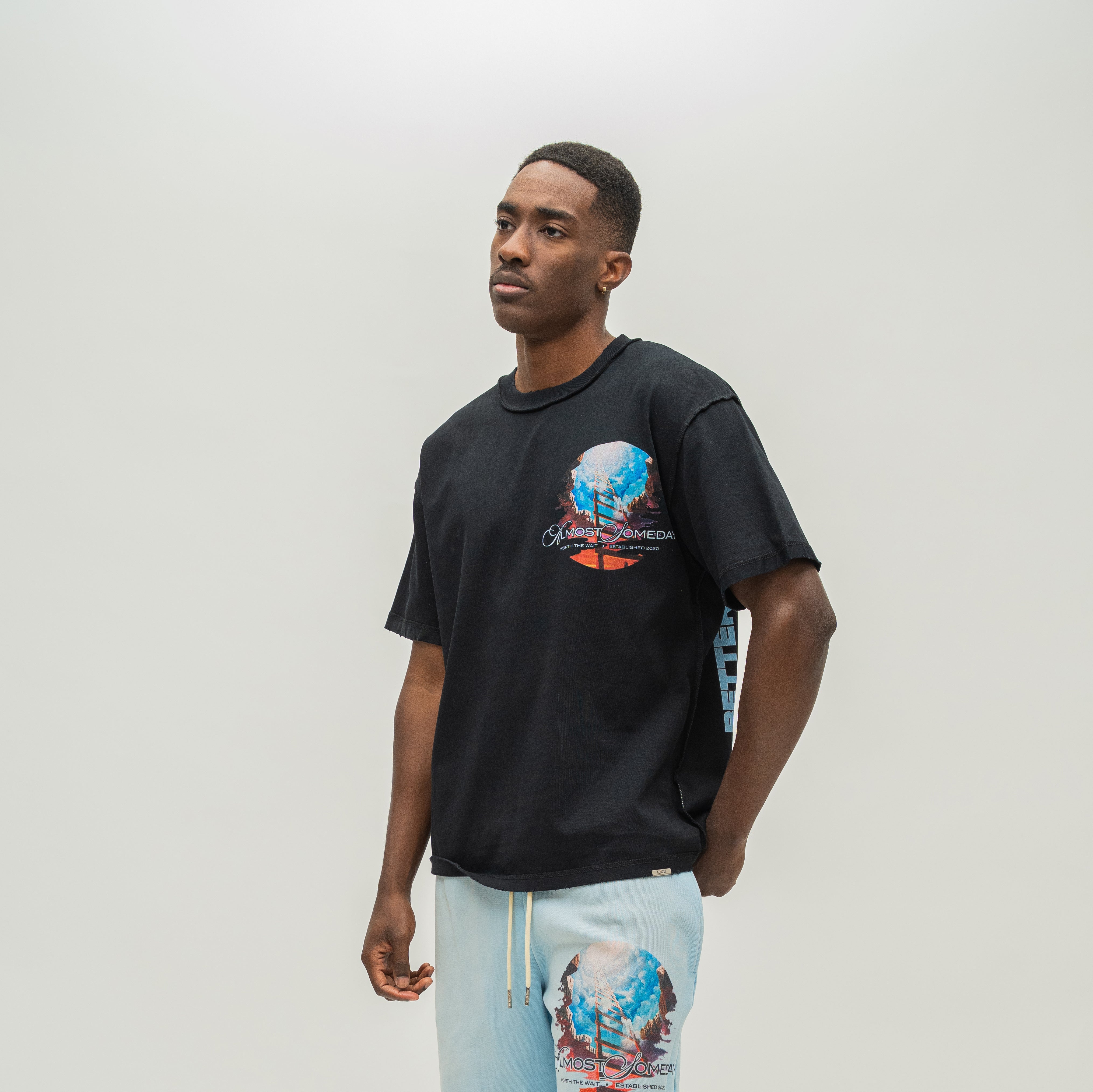 A man wearing a black t-shirt with a colorful graphic on the chest and light blue sweatshorts, standing against a plain light gray background.