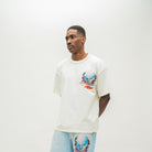 A man wearing a cream-colored oversized t-shirt with a colorful graphic print on the left chest, paired with light blue sweatshorts, standing against a plain off-white studio background.