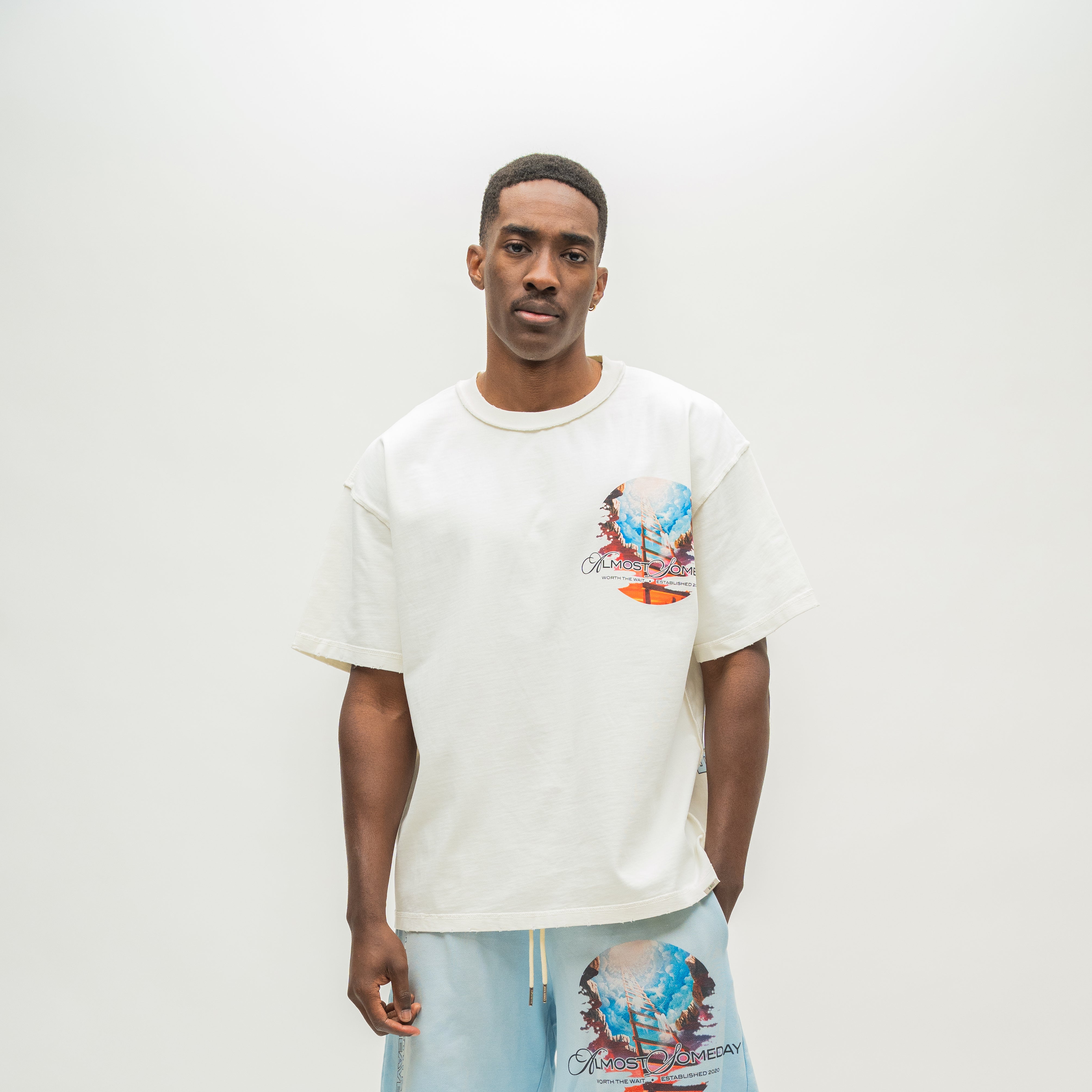 A man wearing an oversized off-white graphic t-shirt and light blue shorts with a matching graphic print, standing against a plain light gray studio background.