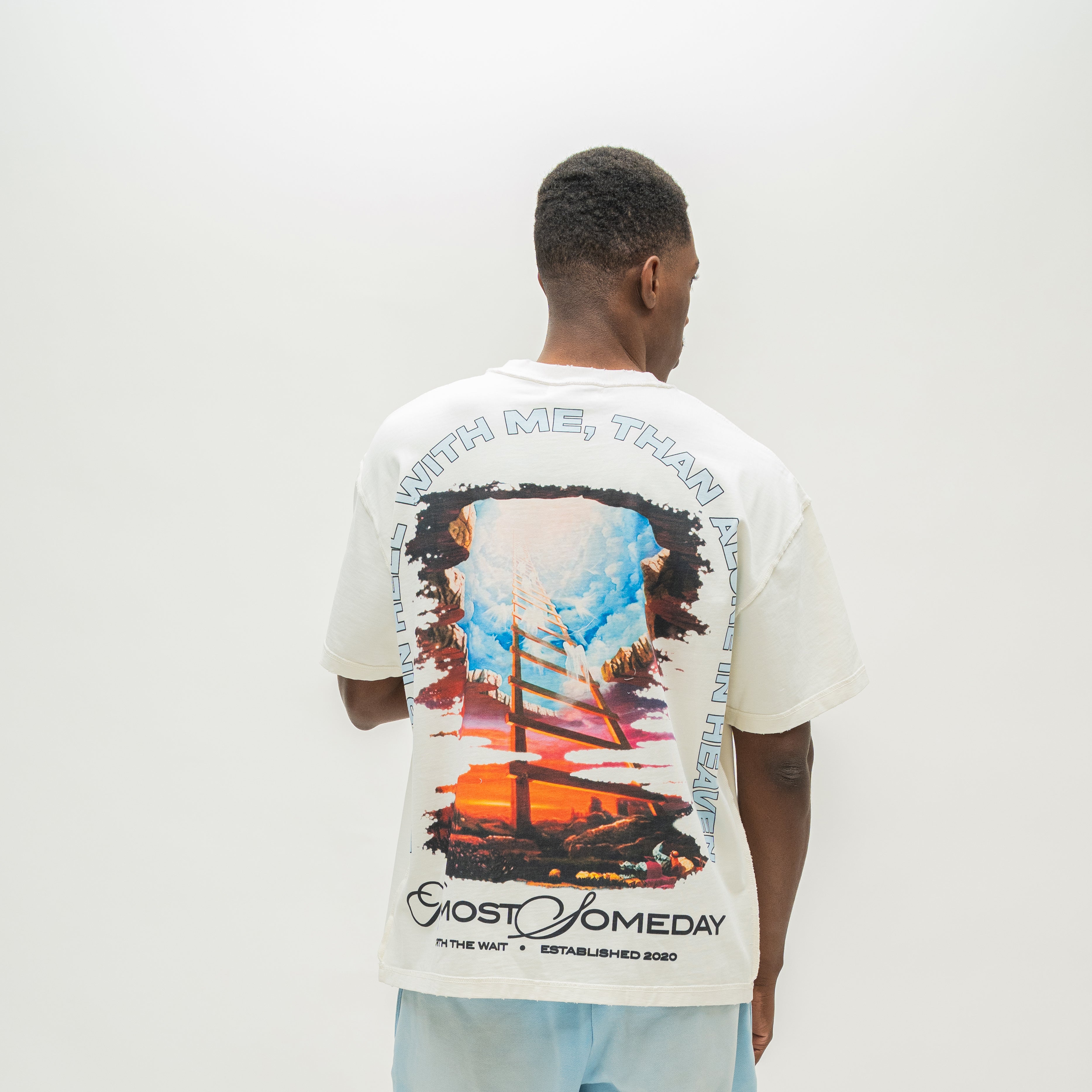 A man viewed from behind wearing a white Almost Someday graphic t-shirt with a large colorful print of a sailing ship and text, paired with light blue sweatshorts against a plain white background.