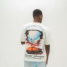 A man viewed from behind wearing a white Almost Someday graphic t-shirt with a large colorful print of a sailing ship and text, paired with light blue sweatshorts against a plain white background.