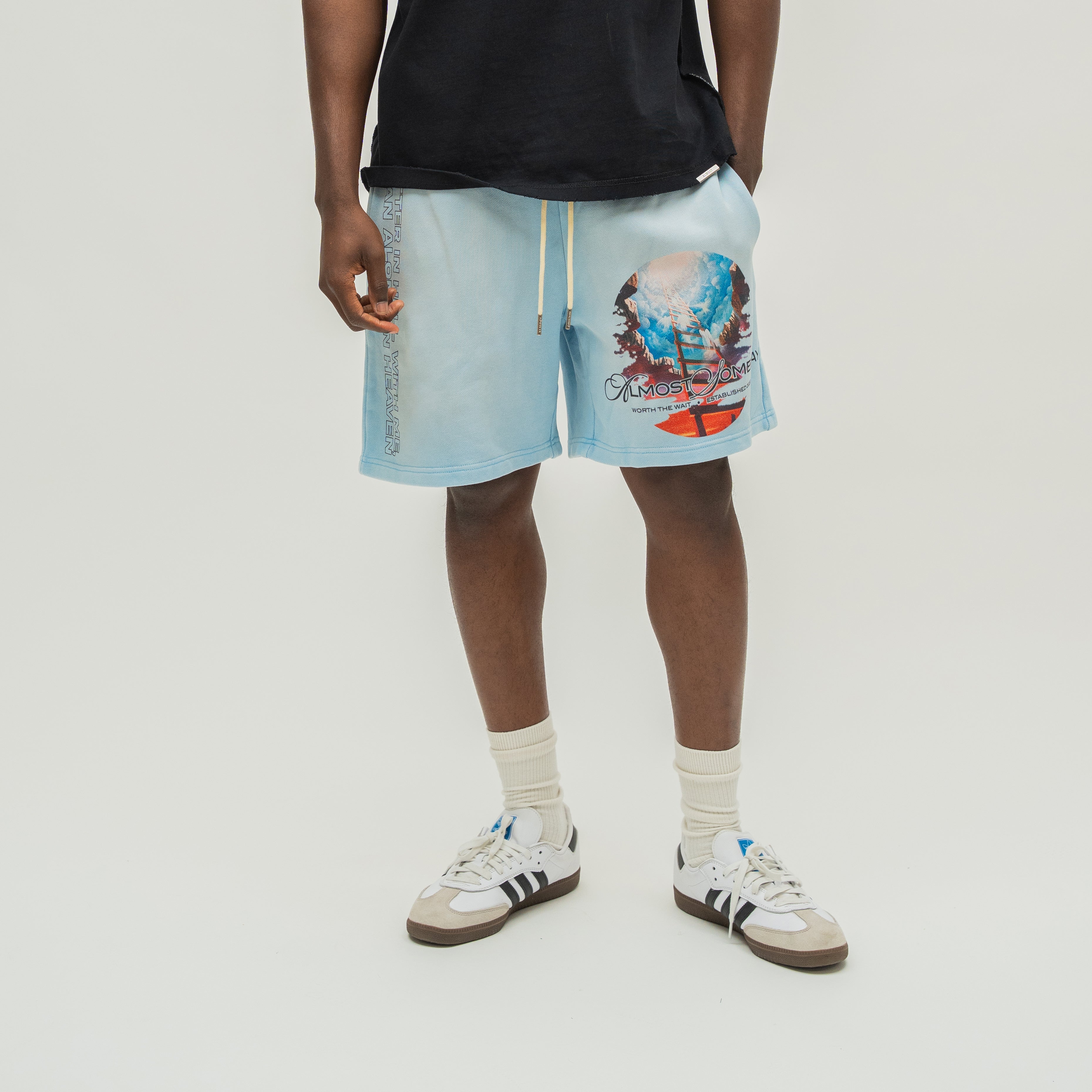 A person wearing light blue drawstring shorts with a colorful graphic print on the left leg, paired with a black t-shirt, white socks, and white sneakers with black stripes, against a light gray studio background.