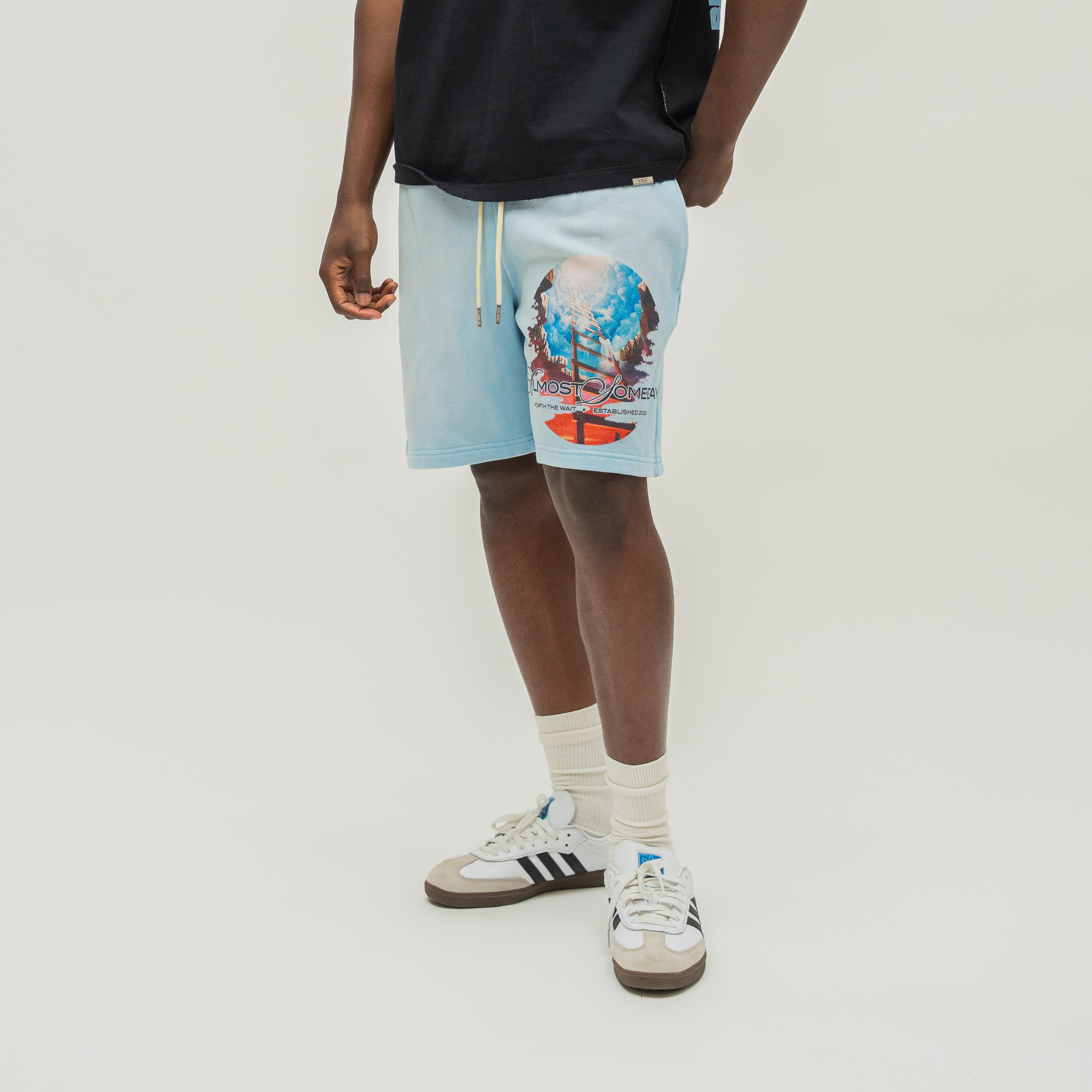 A person wearing light blue athletic shorts with a colorful graphic print on the left leg, paired with a black t-shirt, white socks, and white sneakers with black stripes, against a light gray studio background.