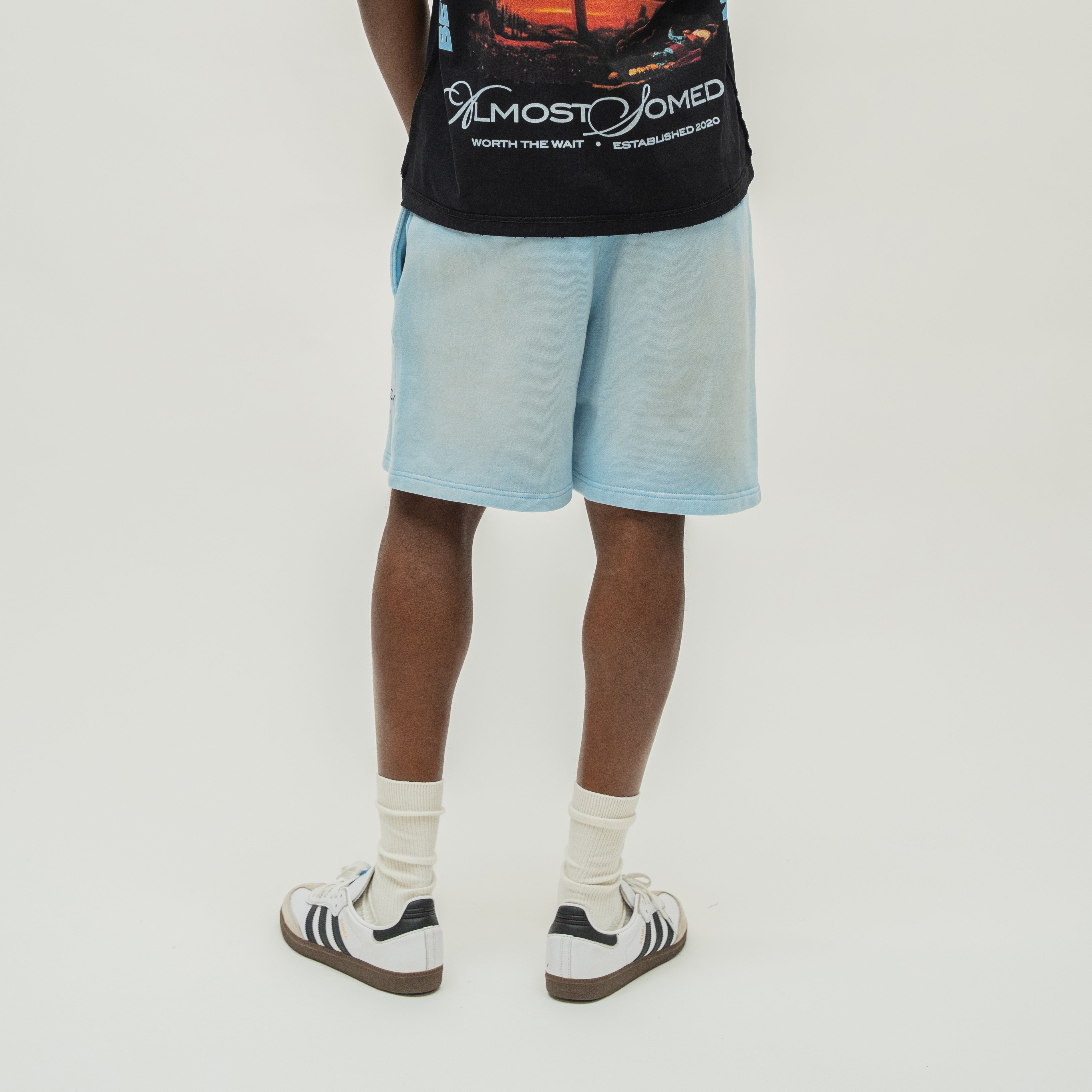 A person shown from the back wearing light blue denim shorts, a black graphic t-shirt, white socks, and white sneakers against a plain light gray studio background.