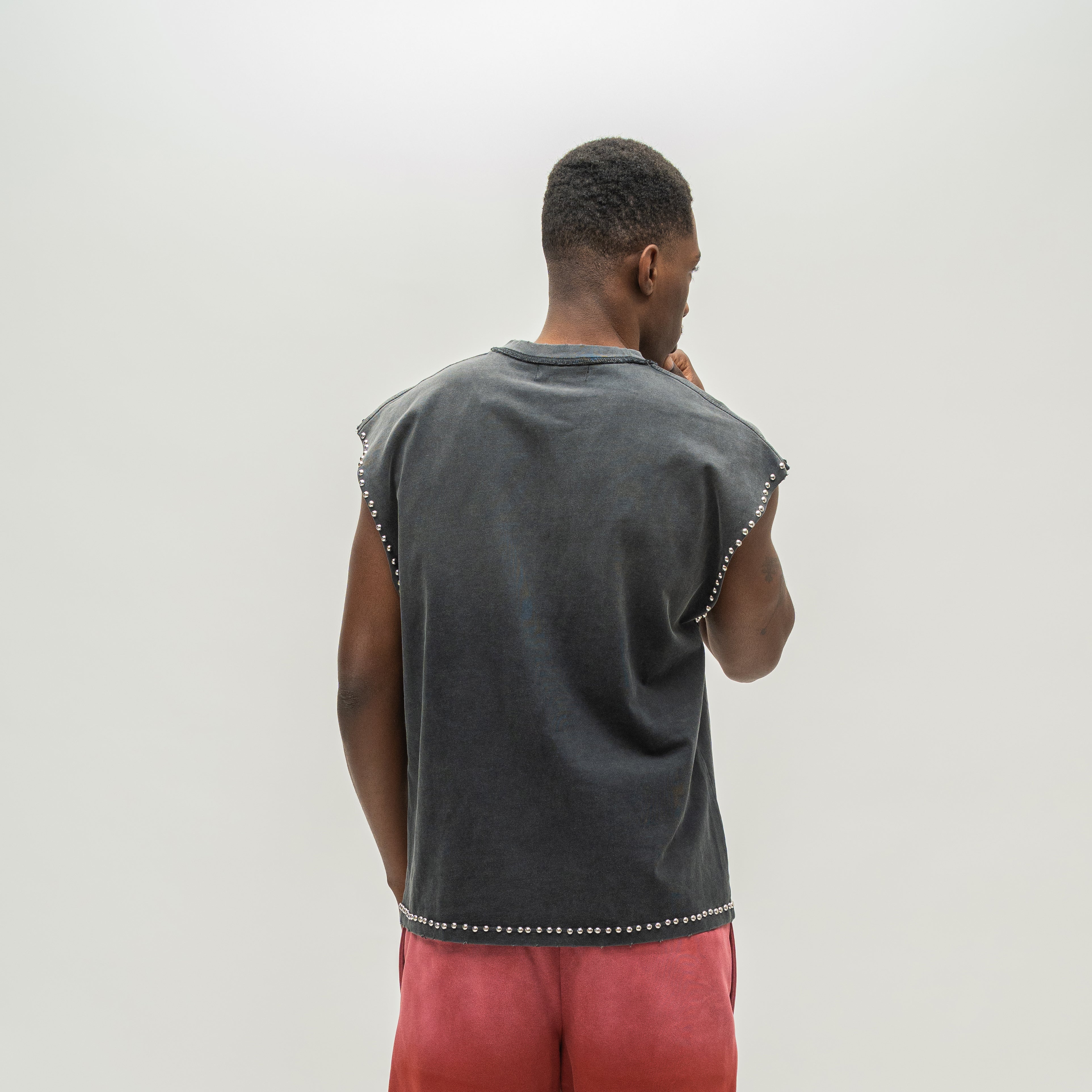 A man viewed from behind wearing a dark gray sleeveless t-shirt with silver stud detailing along the edges, paired with coral pink pants, against a plain light gray background.
