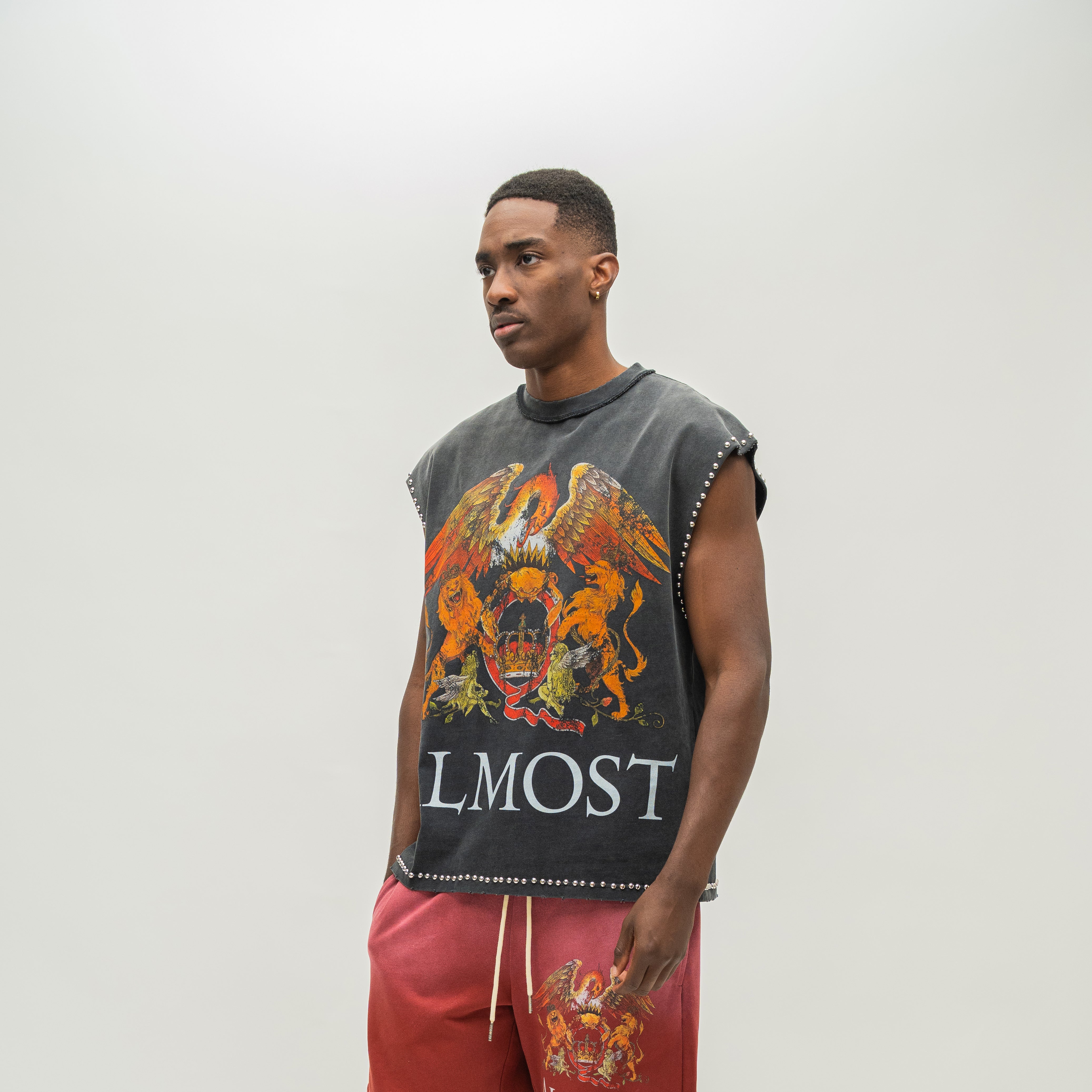 A man modeling a sleeveless black t-shirt with a large orange and gold eagle graphic and the word 'ALMOST' printed on the front, paired with red sweatpants against a plain light gray background.