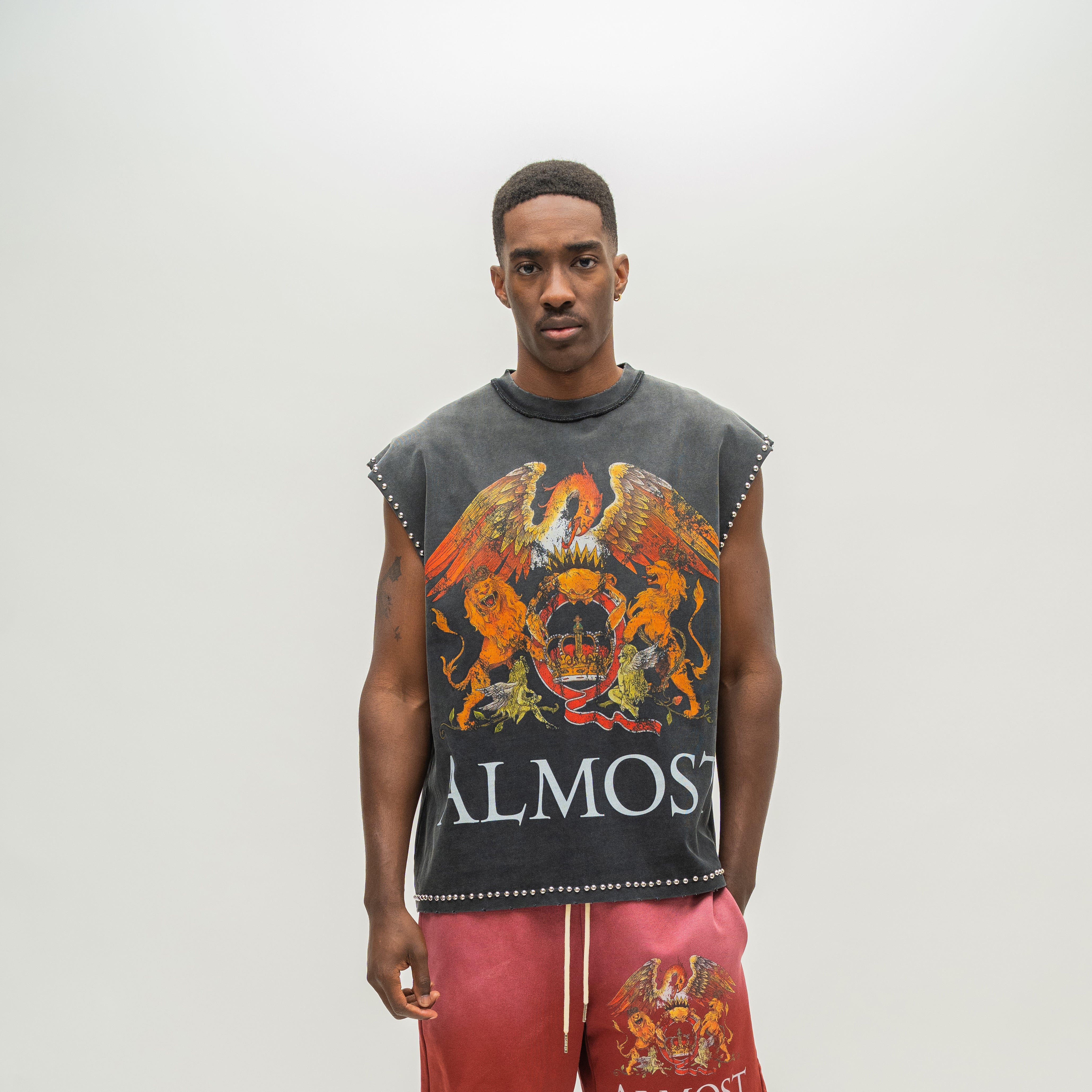 A man modeling a sleeveless dark gray distressed graphic top with a large multicolor eagle and skull print and the word 'ALMOST' on the front, paired with pink drawstring shorts against a light gray studio background.