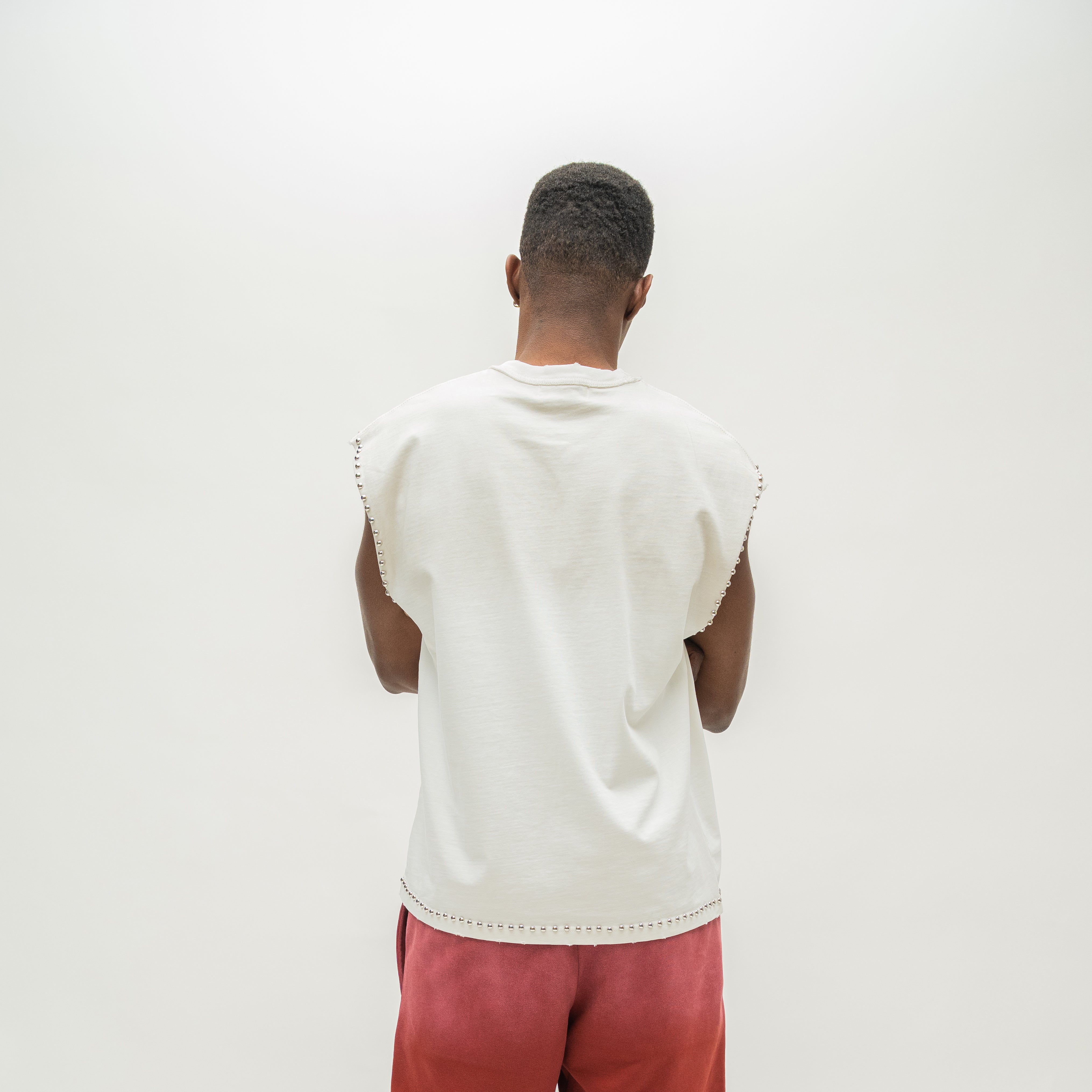 A man viewed from behind wearing a sleeveless white t-shirt with scalloped edges and pink pants, standing against a plain white background.