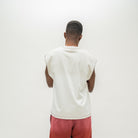 A man viewed from behind wearing a sleeveless white t-shirt with scalloped edges and pink pants, standing against a plain white background.