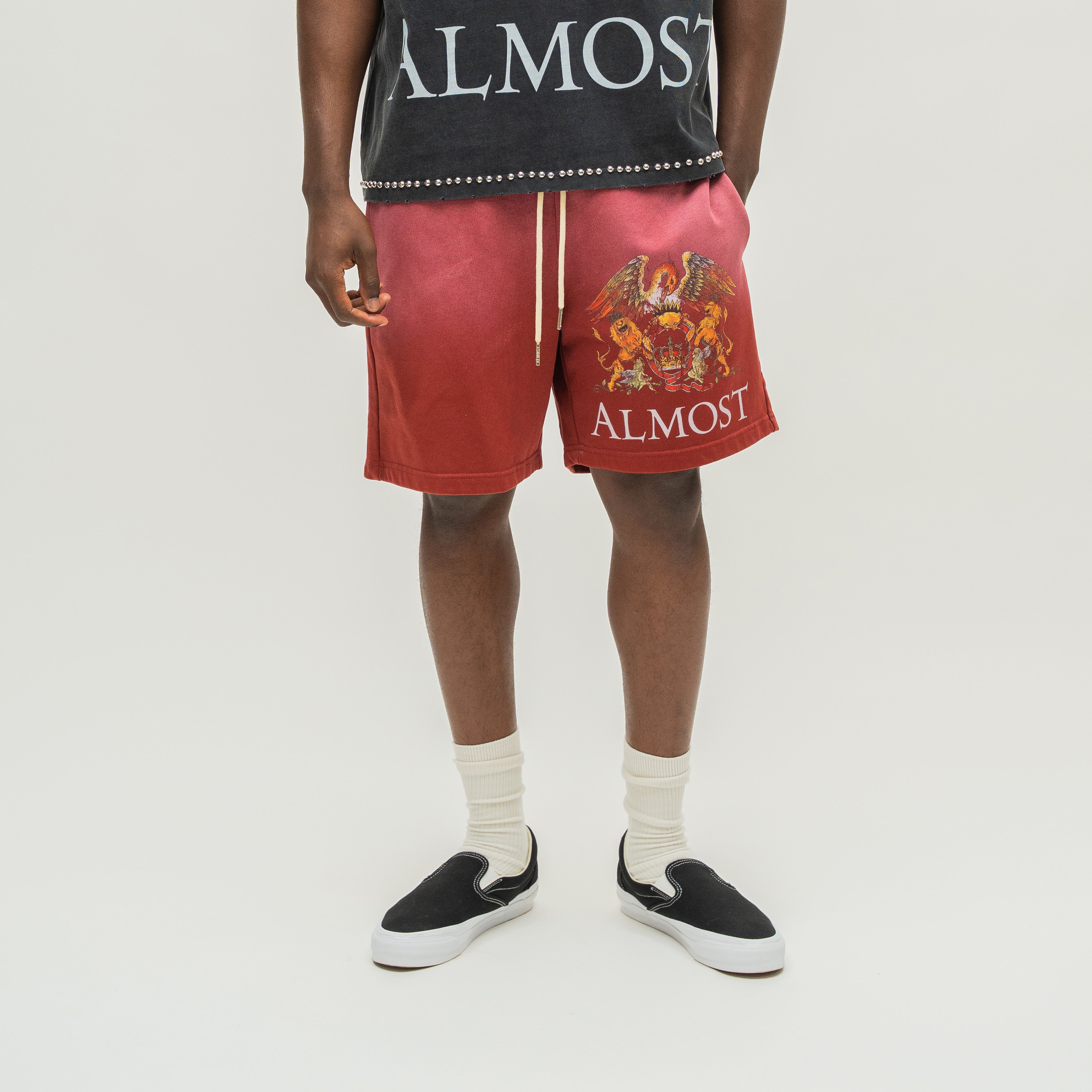 A model is shown from the waist down wearing red ALMOST brand drawstring shorts with a large multicolor crest graphic and white 'ALMOST' text on the left leg, paired with a black graphic t-shirt, white socks, and black slip-on sneakers against a light gray background.