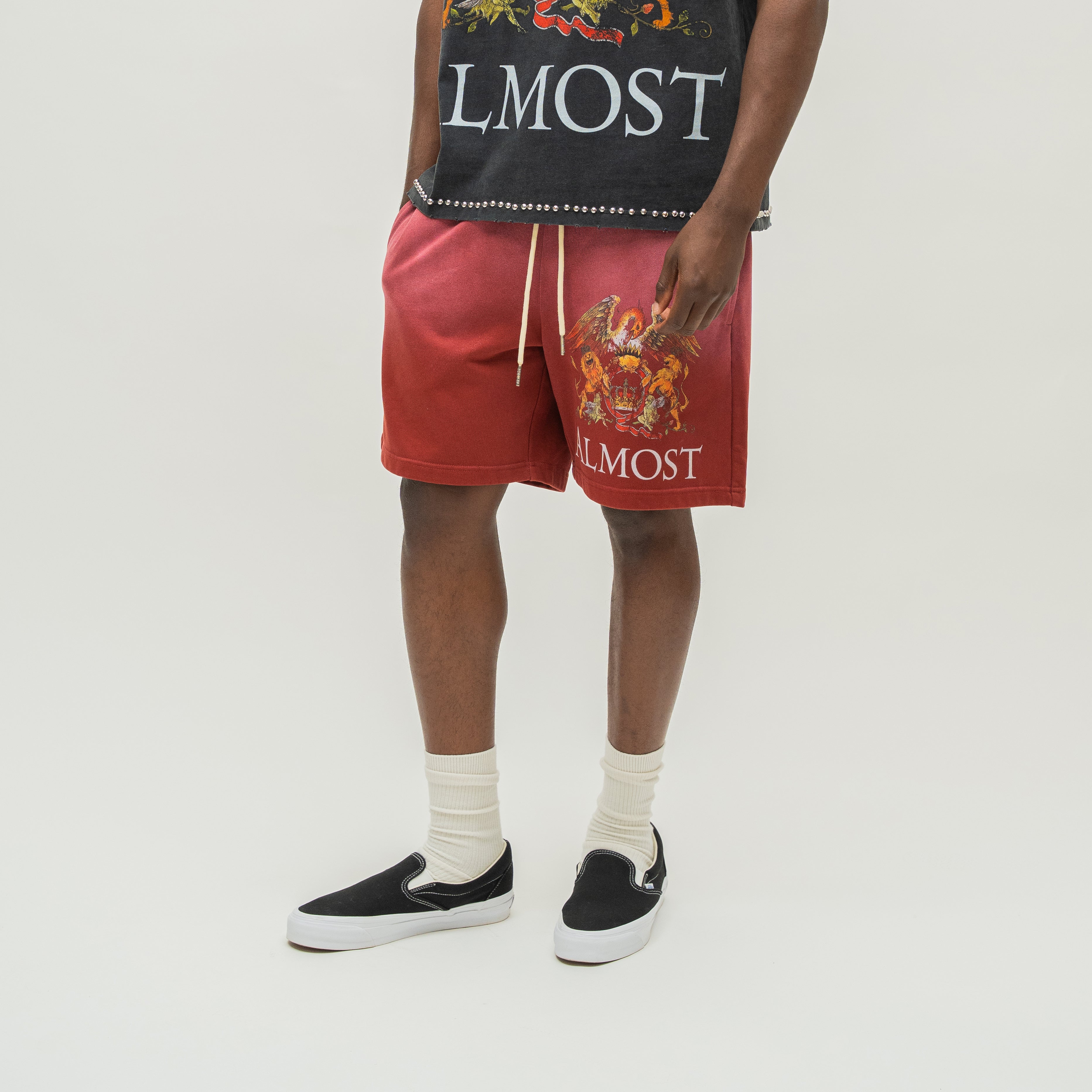 A model is shown from the waist down wearing red drawstring shorts with a large multicolor graphic and the word 'ALMOST' printed on the leg, paired with a black graphic tank top, white socks, and black slip-on shoes against a light gray studio background.