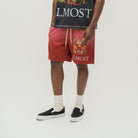 A model is shown from the waist down wearing red drawstring shorts with a large multicolor graphic and the word 'ALMOST' printed on the leg, paired with a black graphic tank top, white socks, and black slip-on shoes against a light gray studio background.