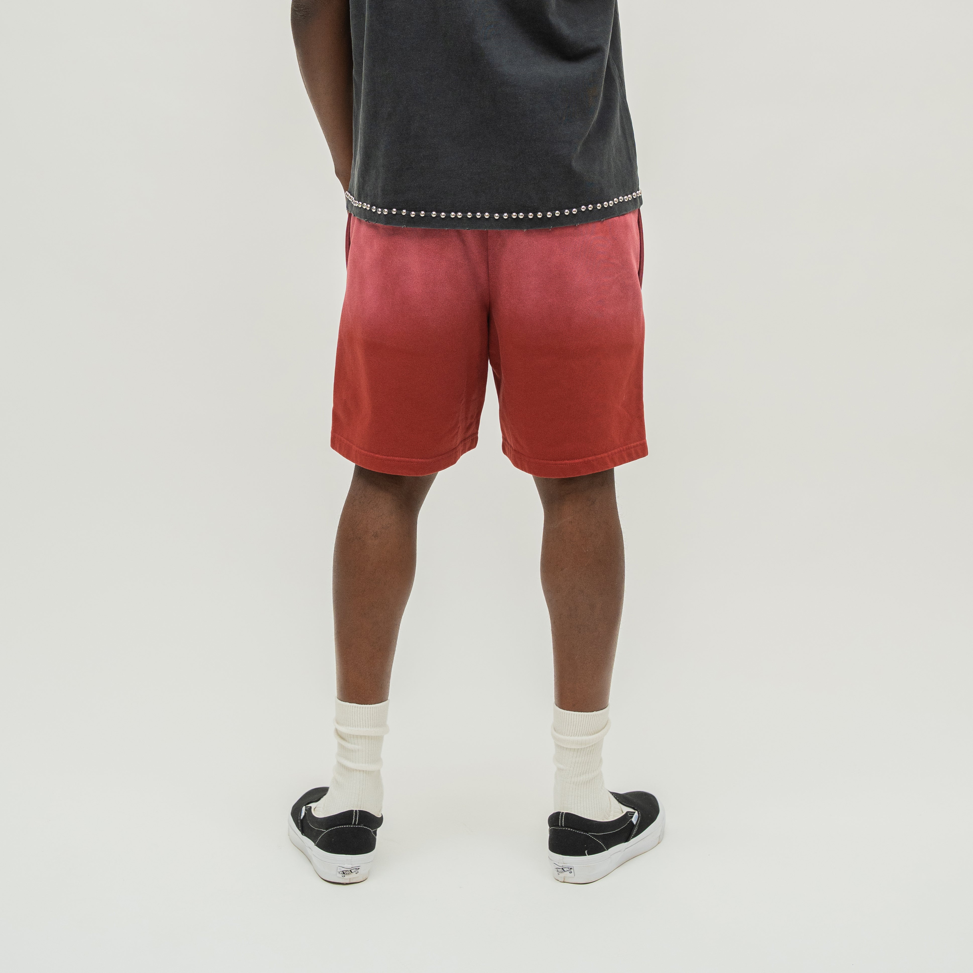A person shown from the back wearing red gradient tie-dye shorts, a dark gray t-shirt, white socks, and black slip-on sneakers against a light gray studio background.