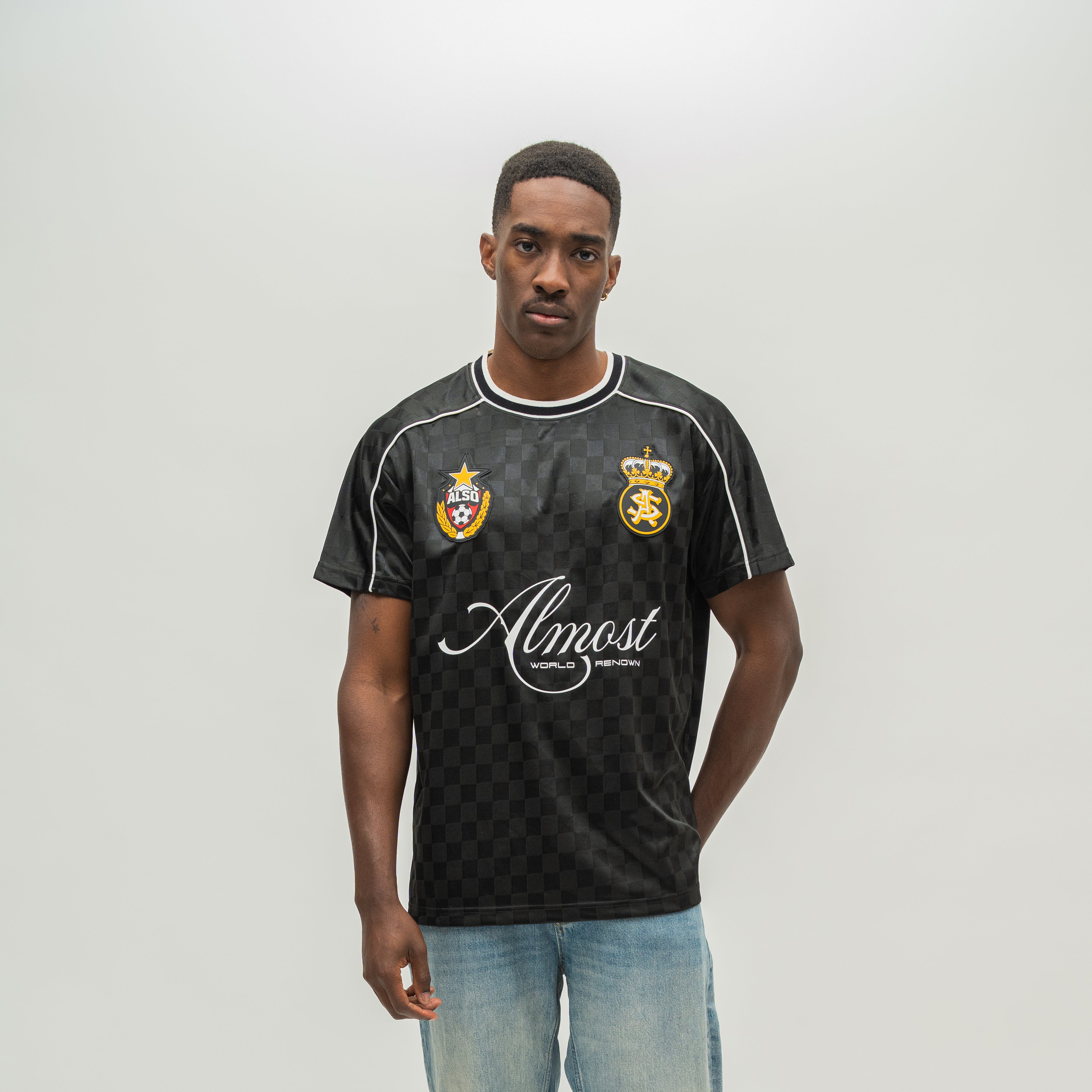 A man modeling a black checkered short-sleeve soccer jersey with white piping, white script text reading 'Almost Someday', and two gold and blue club crests on the chest, paired with light blue jeans against a light gray studio background.