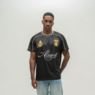 A man modeling a black checkered short-sleeve soccer jersey with white piping, white script text reading 'Almost Someday', and two gold and blue club crests on the chest, paired with light blue jeans against a light gray studio background.