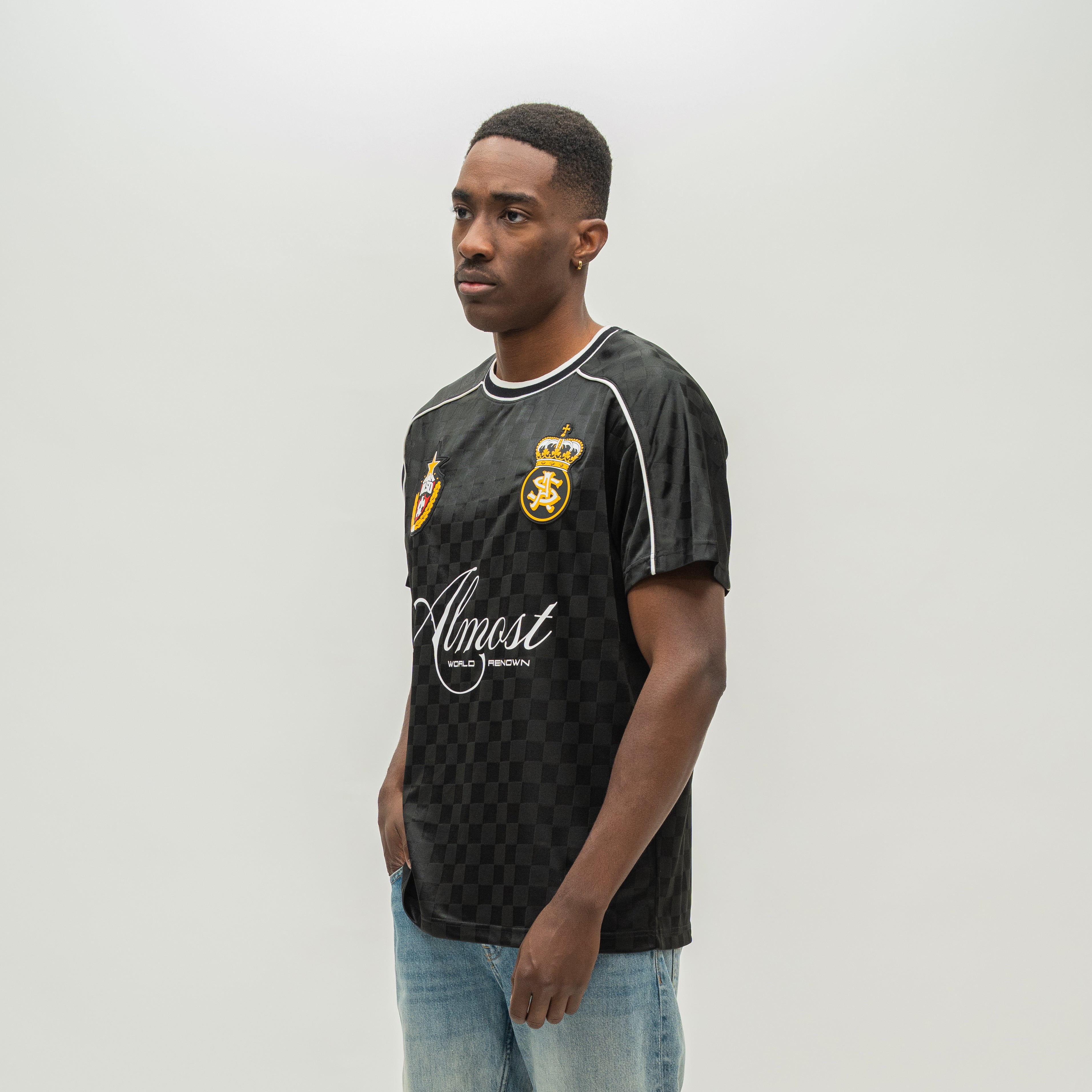 A man wearing a black and dark gray checkered t-shirt with white piping, a gold crest, and a white 'Almost Someday' logo, paired with light blue jeans, standing against a light gray studio background.